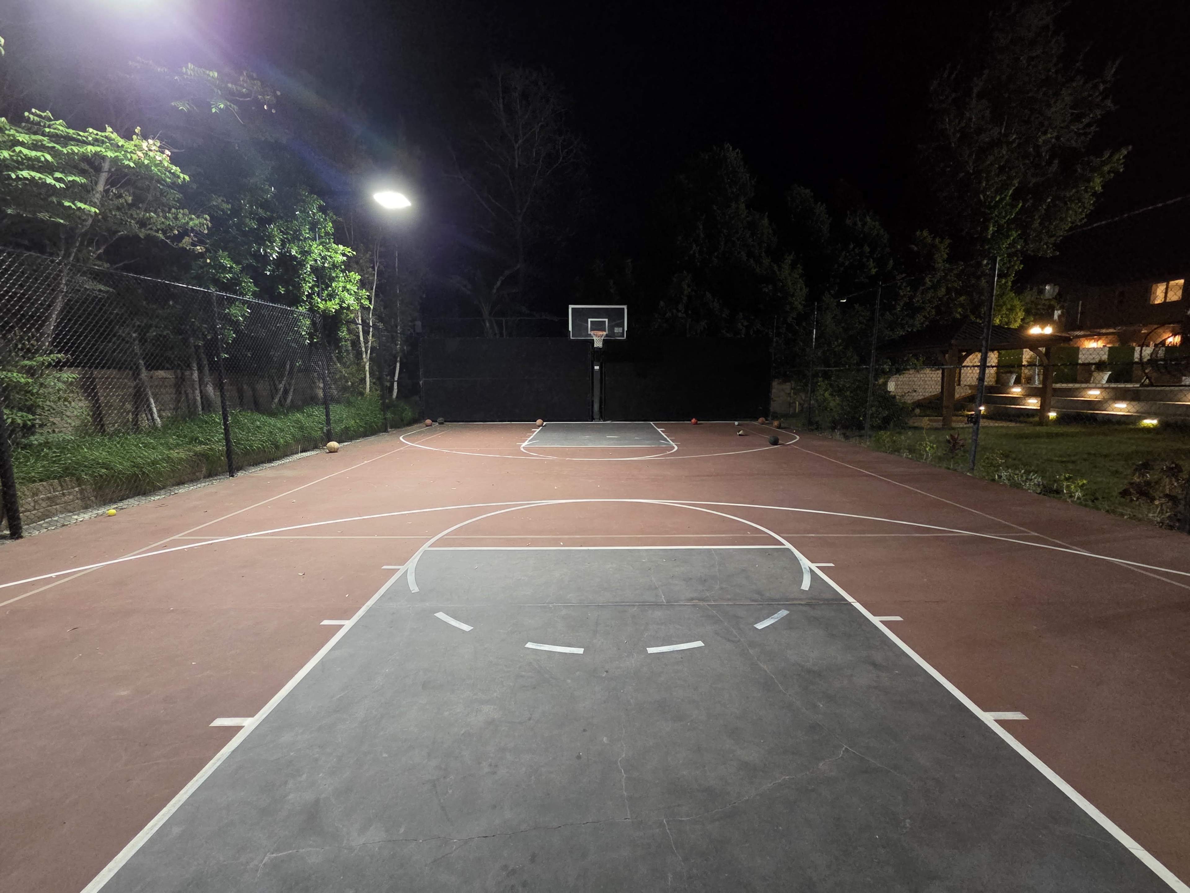 The image shows a basketball court at night, illuminated by a nearby light, with a clear view of the hoop and markings on the court.