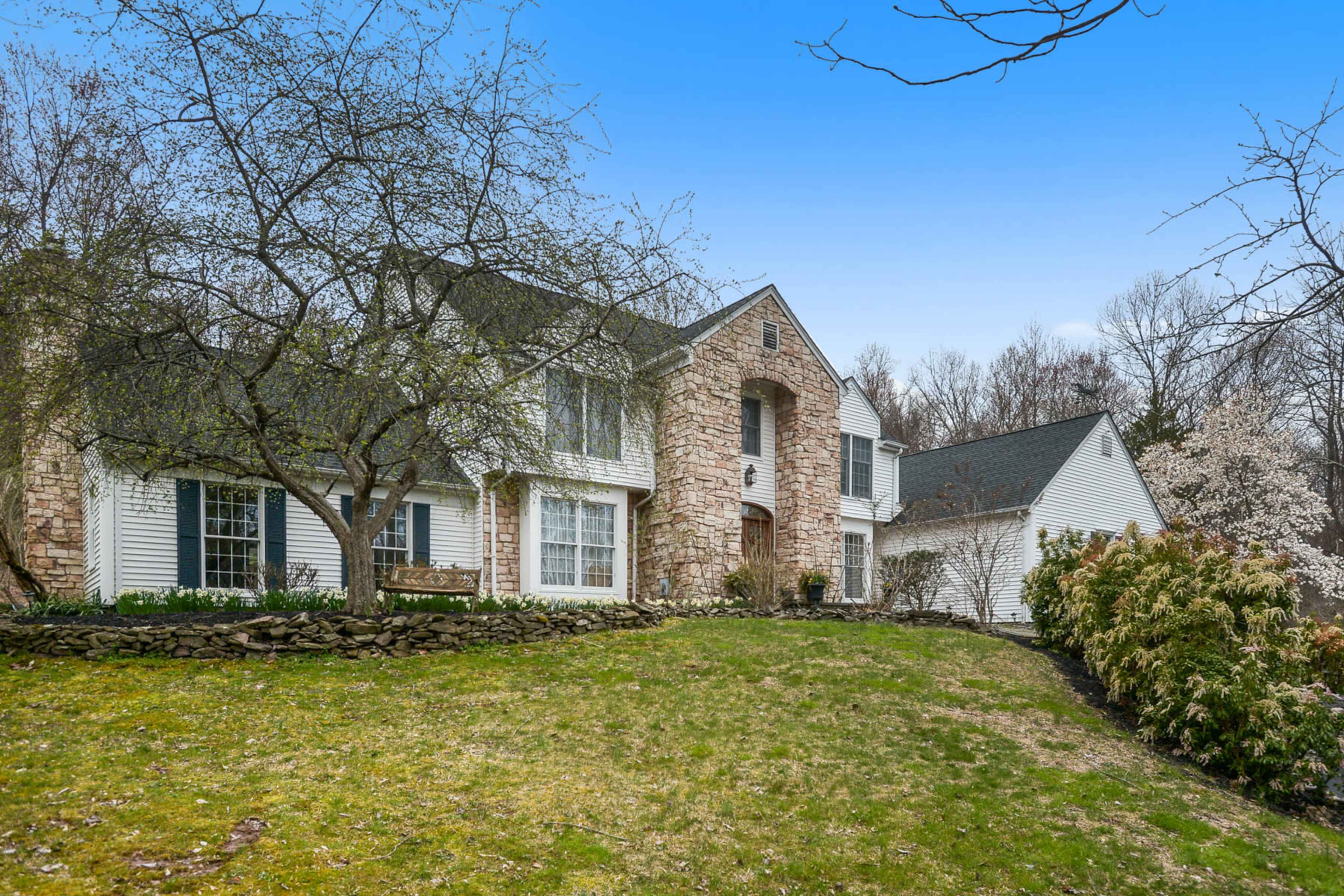 A large stone and siding house is set on a grassy lawn, surrounded by trees and shrubs.