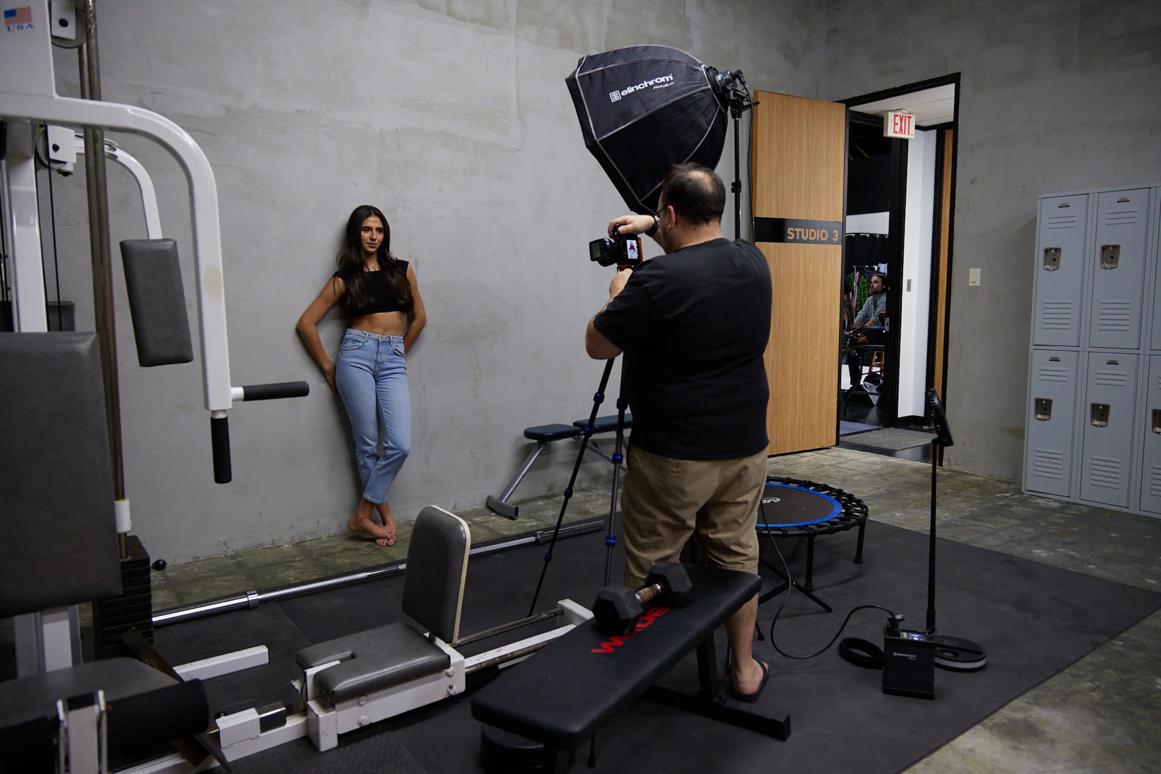 A photographer is capturing images of a model posing against a concrete wall in a studio that features various gym equipment.