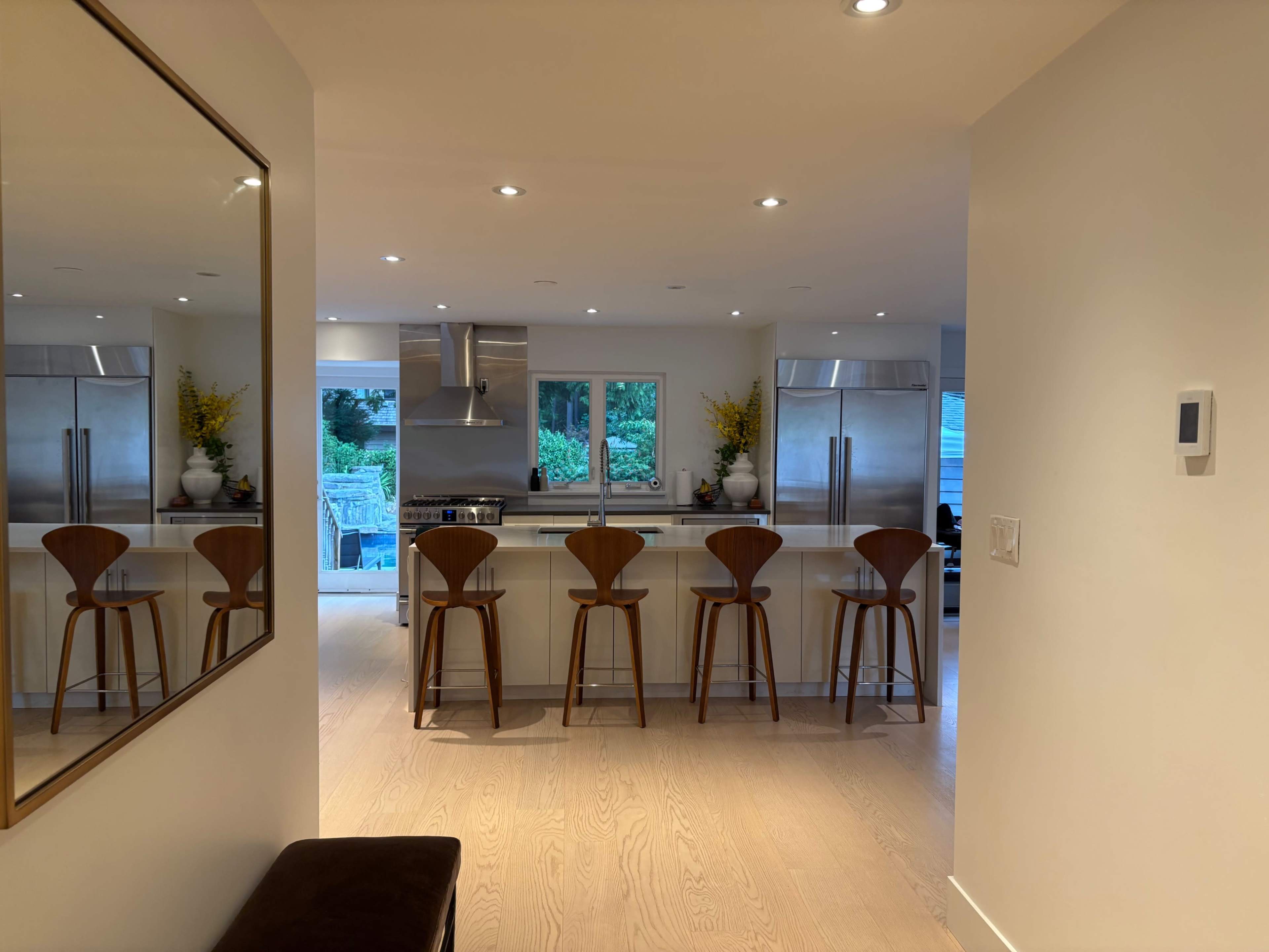 A modern kitchen with stainless steel appliances and four wooden bar stools lined up at a countertop, viewed from a hallway with a mirror on the wall.