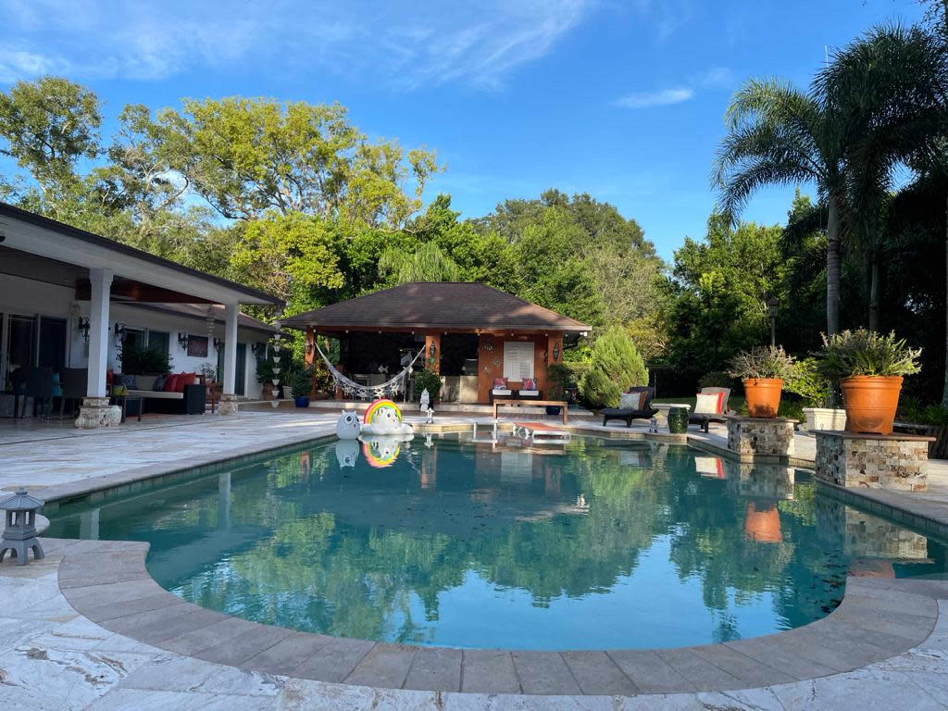 The image shows a residential backyard with a large swimming pool surrounded by stone borders, a gazebo, potted plants, and sun loungers.