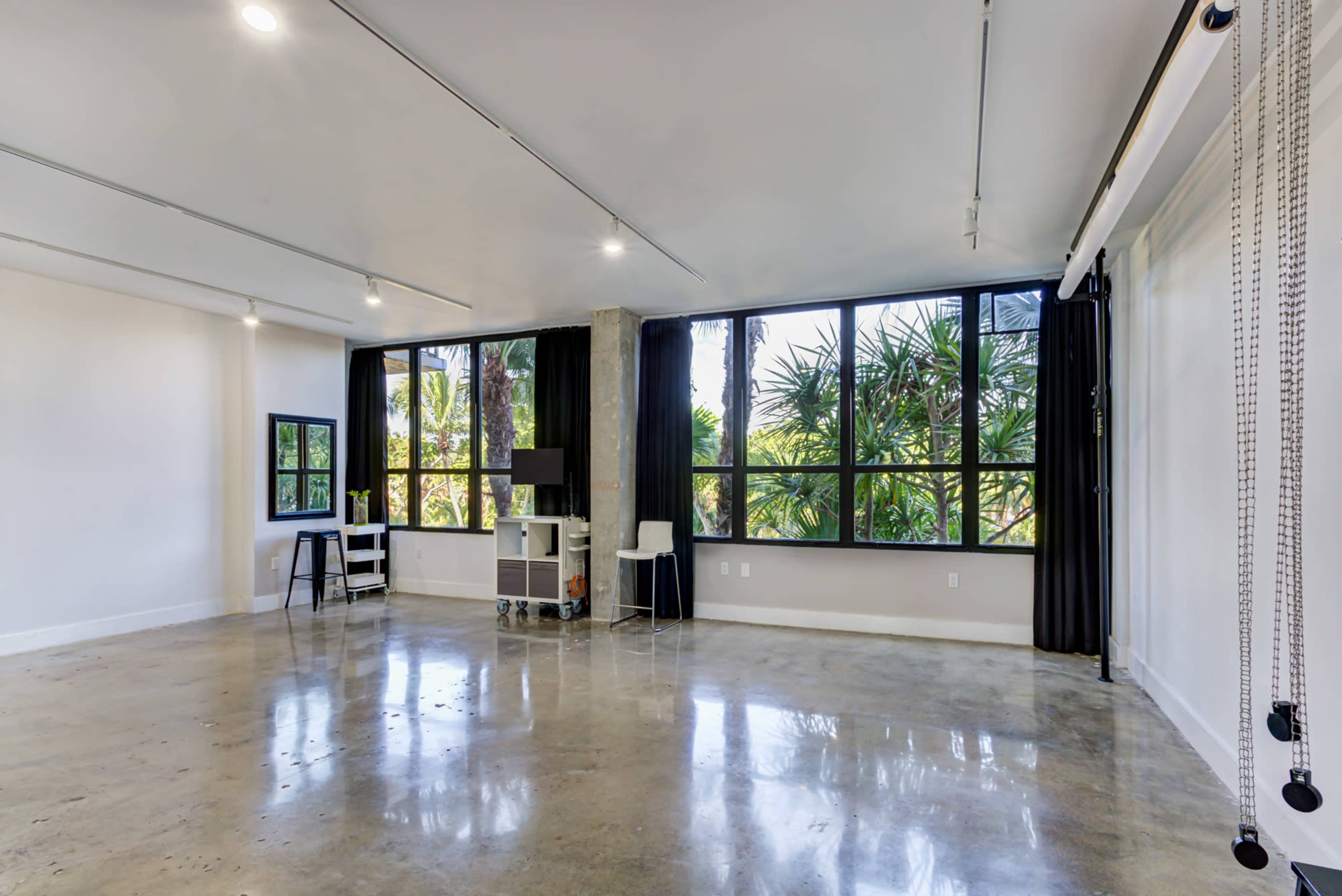 A spacious room with polished concrete floors, large windows overlooking greenery, and minimal furnishings including a small desk and a bar stool.