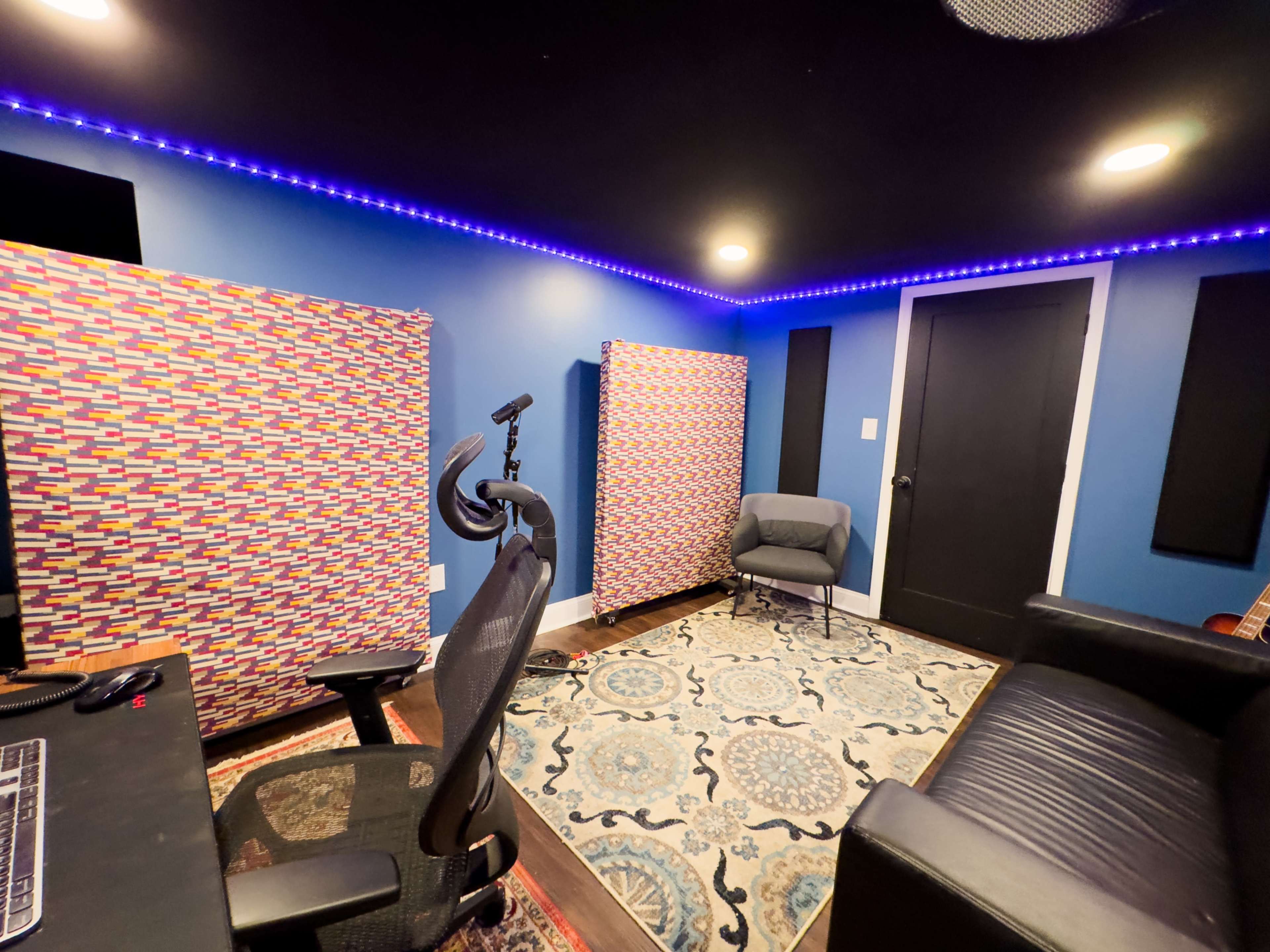 A recording studio with colorful acoustic panels, a desk with a computer, a black leather sofa, and ambient lighting.