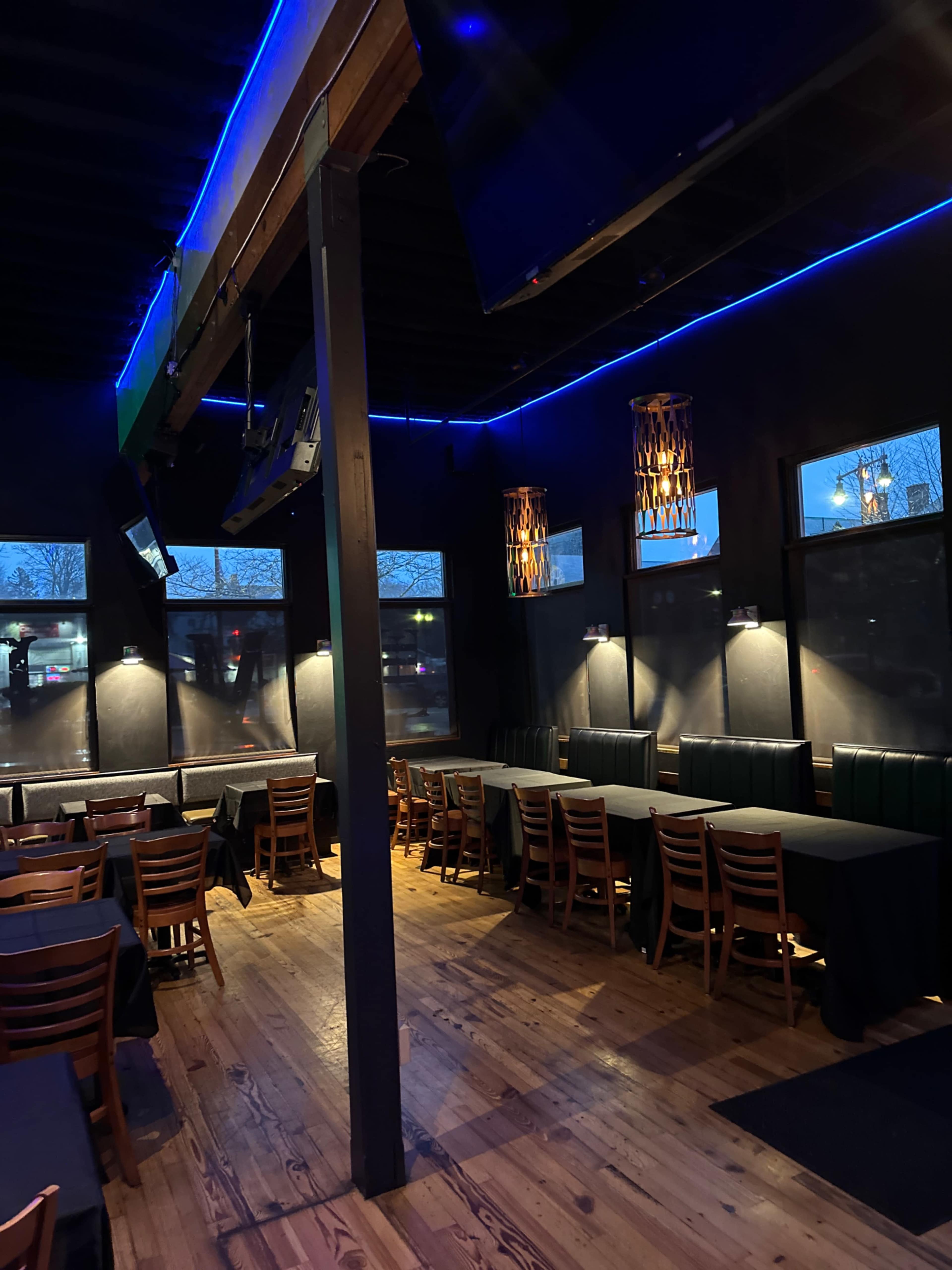 An empty restaurant interior with wooden flooring, dark walls, and several tables arranged with black tablecloths under dim lighting.