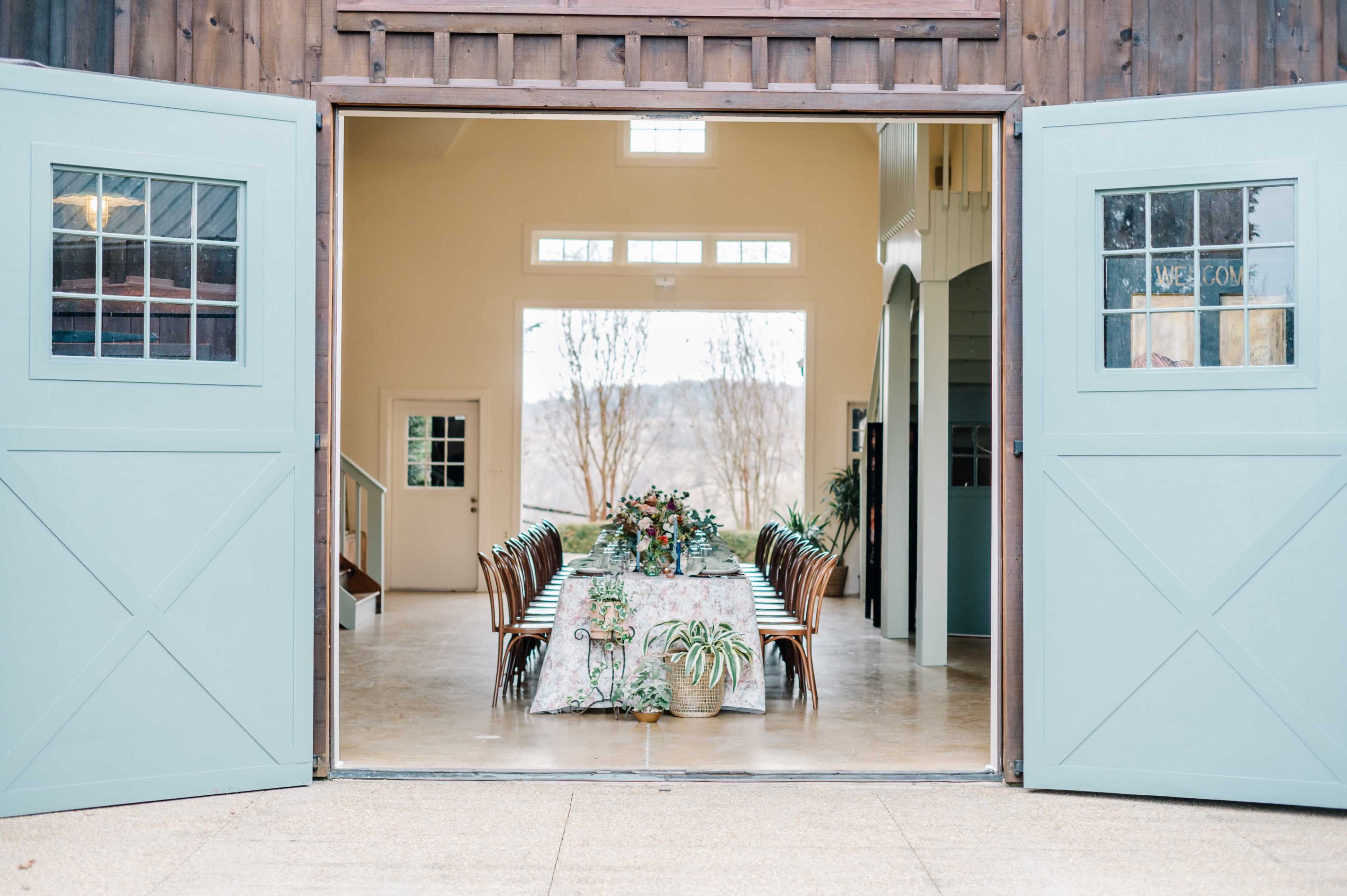 A long dining table is set with decorations inside a spacious barn, visible through open double doors.