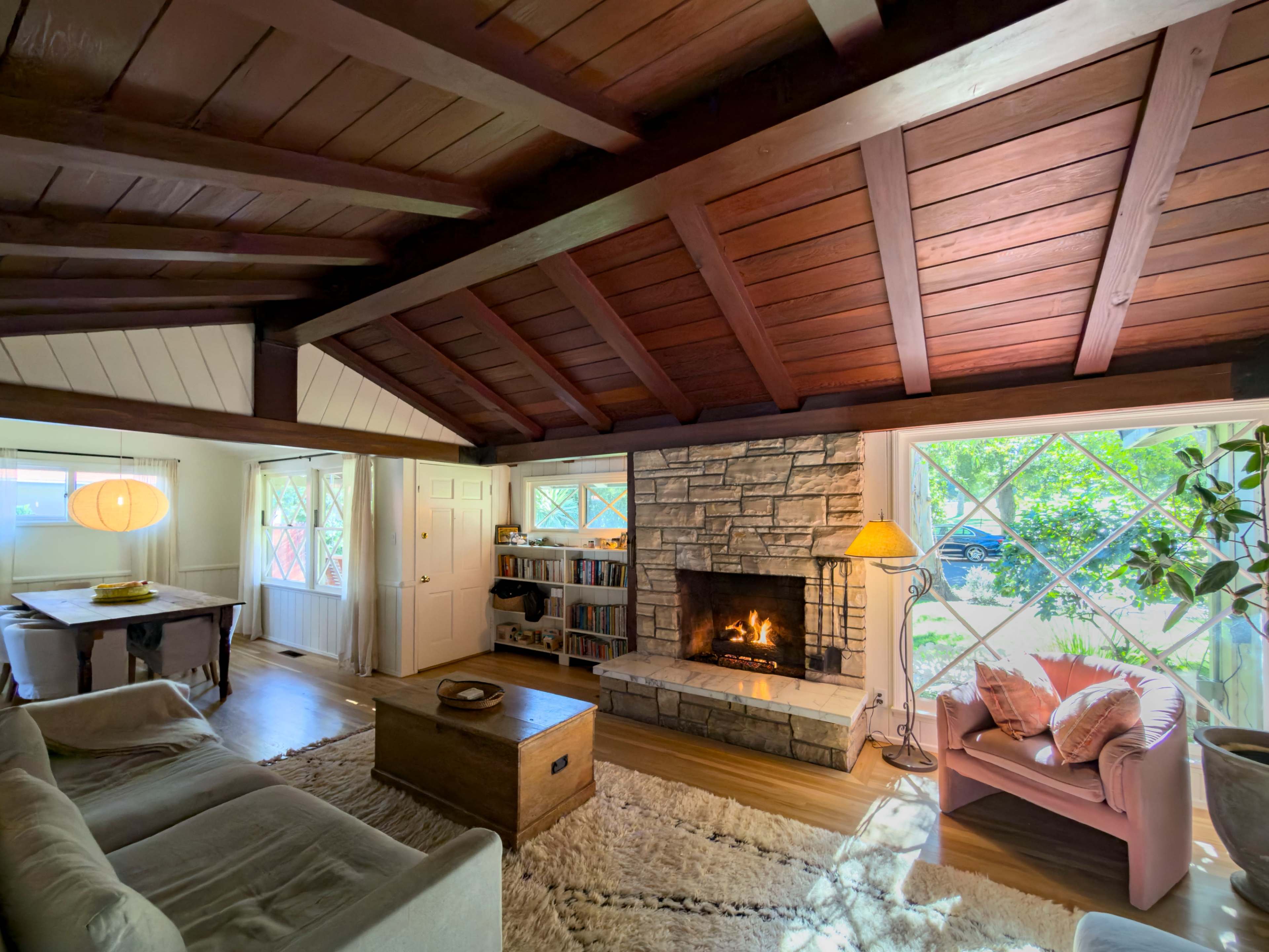 The image shows a cozy living room with a wooden ceiling, a stone fireplace, a light-colored sofa, a wooden coffee table, and a pink armchair near large windows that allow natural light in.
