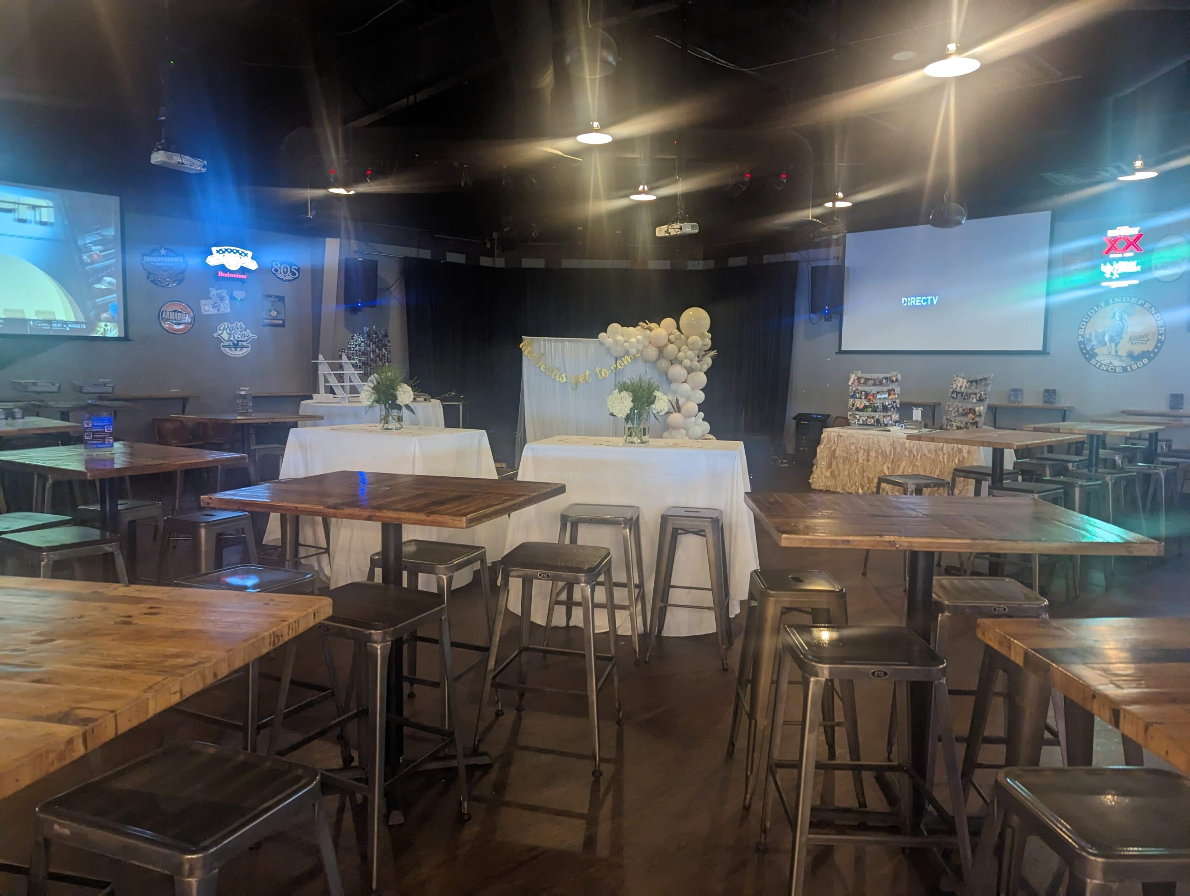A spacious event venue with several wooden tables, a decorative setup at the front, and projection screens on the walls.