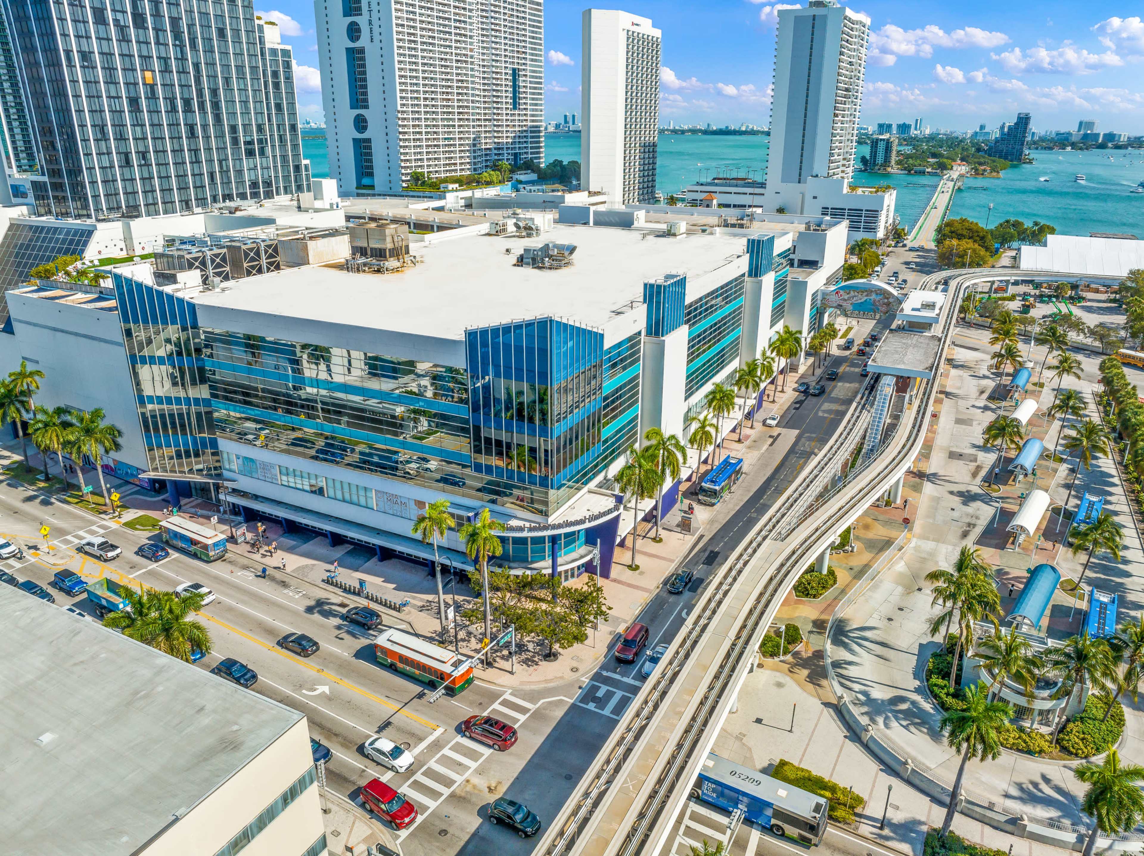 An aerial view of a modern building with large glass windows located near a waterfront, surrounded by palm trees and featuring a transit station with a monorail system.