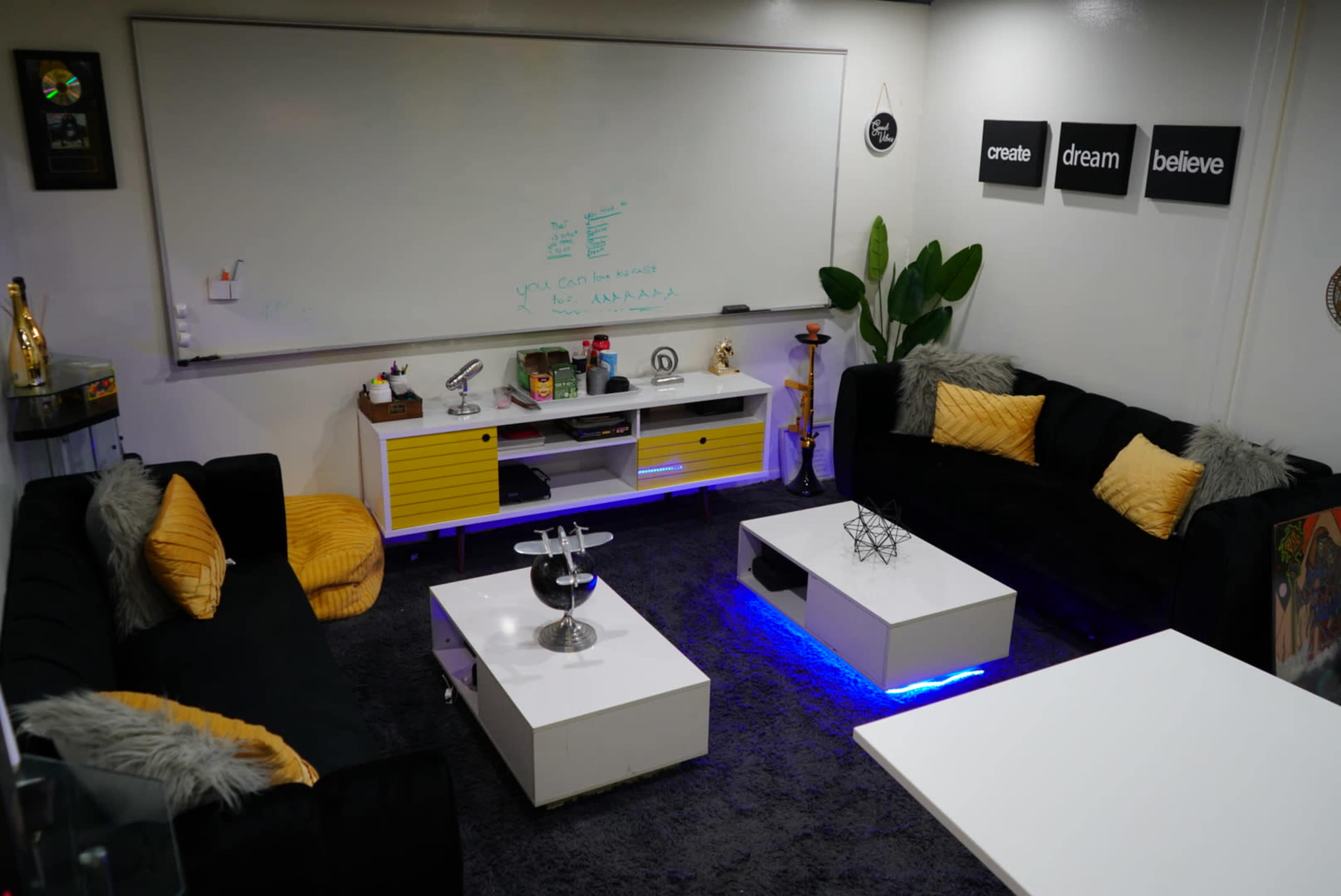 A modern lounge area featuring black sofas with yellow cushions, a low white coffee table with blue lighting, and a whiteboard on the wall surrounded by decorative elements.