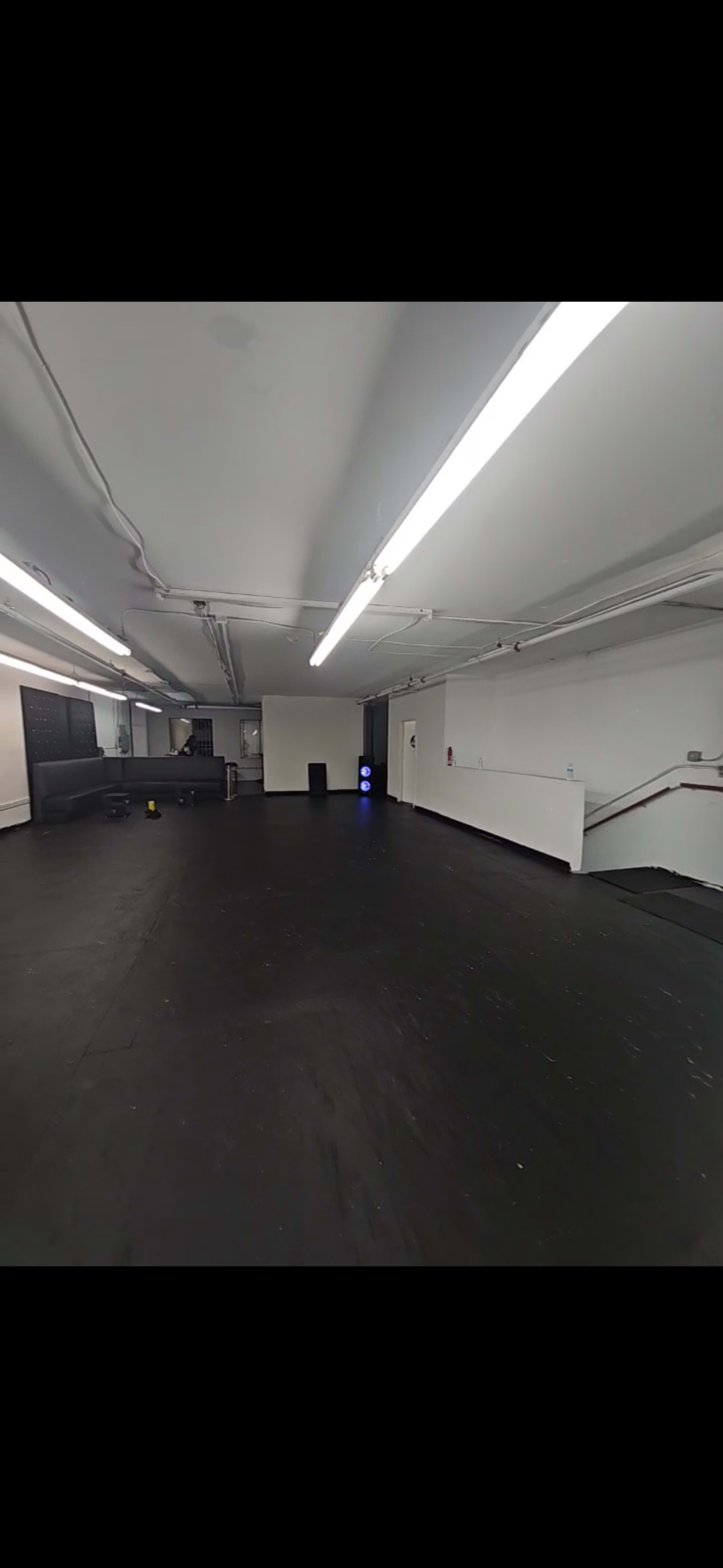 The image shows a spacious, empty room with black flooring, bright overhead lights, and minimal furnishings along the walls.