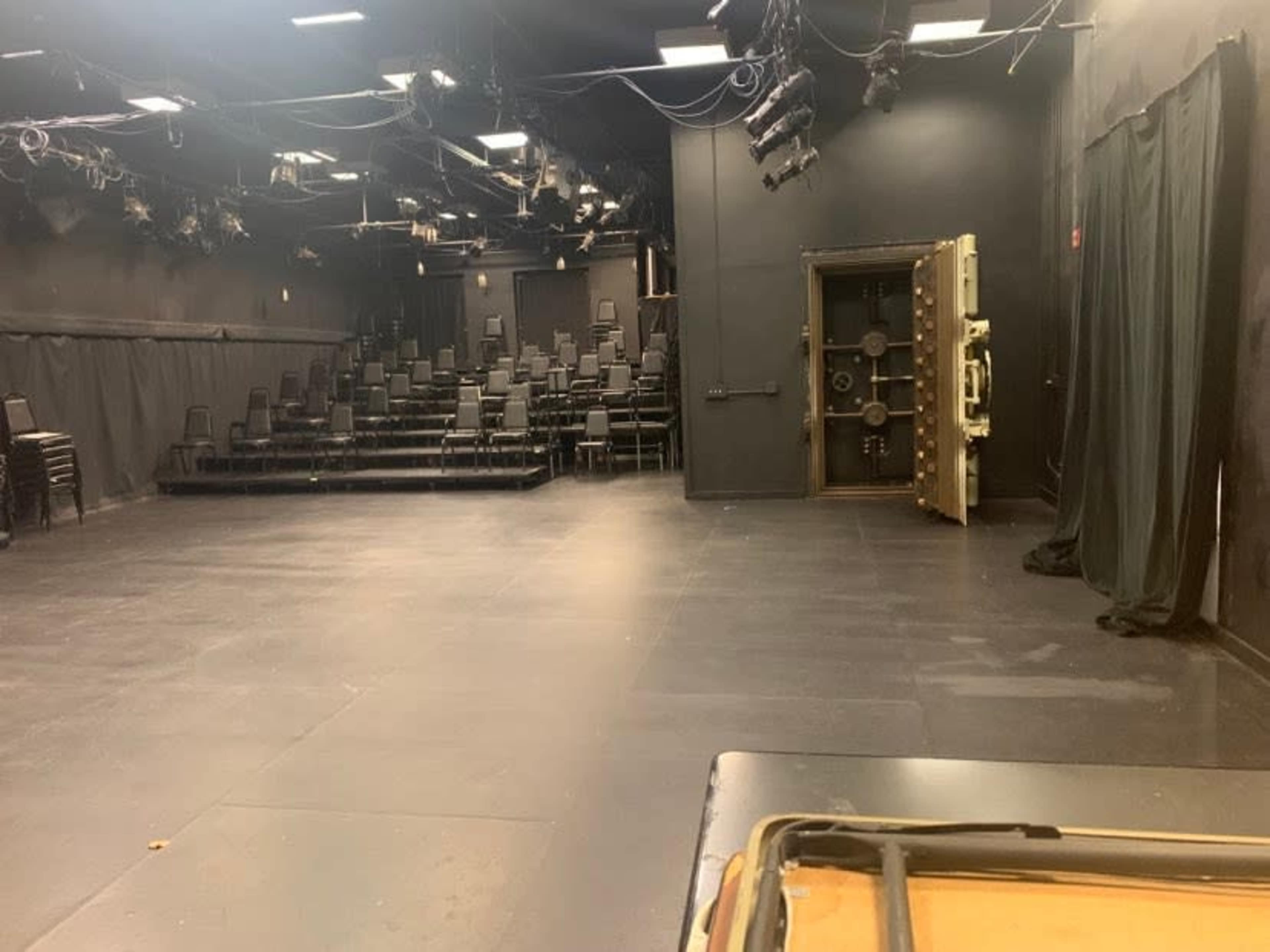 The image shows a black-box theater with rows of stacked chairs, a safe door on the right, and dark curtains in the background.
