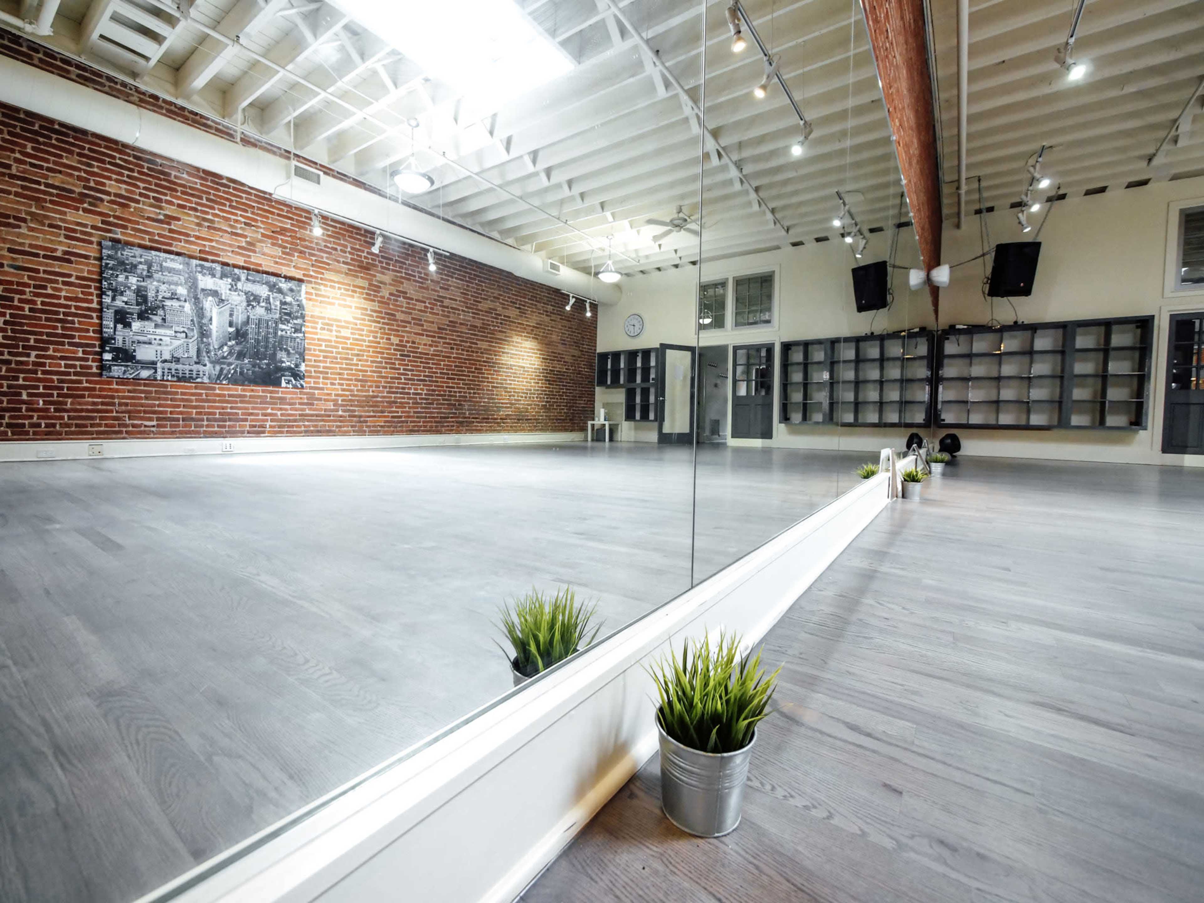 The image shows a spacious studio with a wooden floor, exposed brick walls, a large mirror, and a small potted plant along the edge.