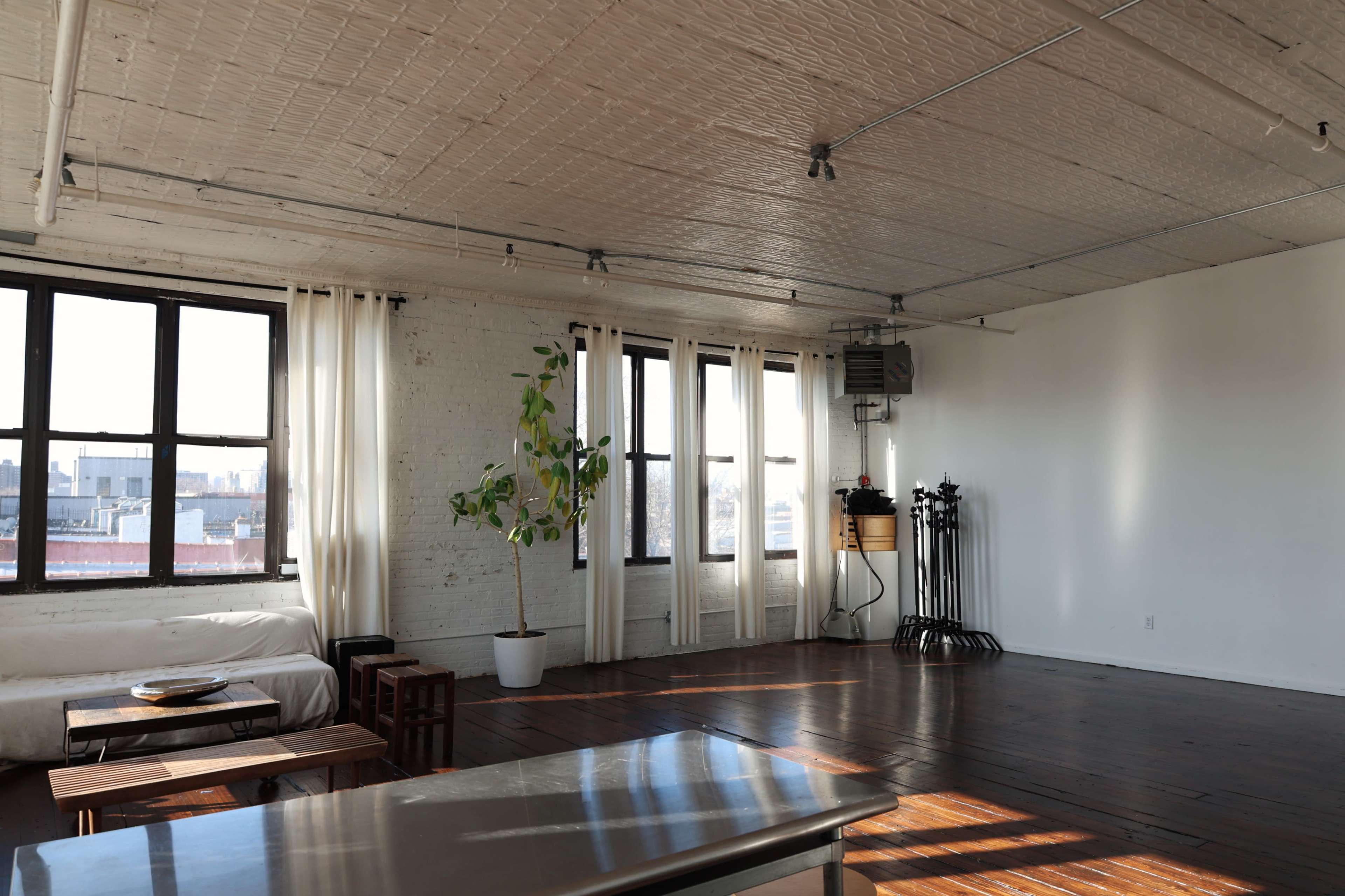 The image depicts a spacious, minimalist studio apartment with large windows, wooden flooring, and a potted plant in the corner.