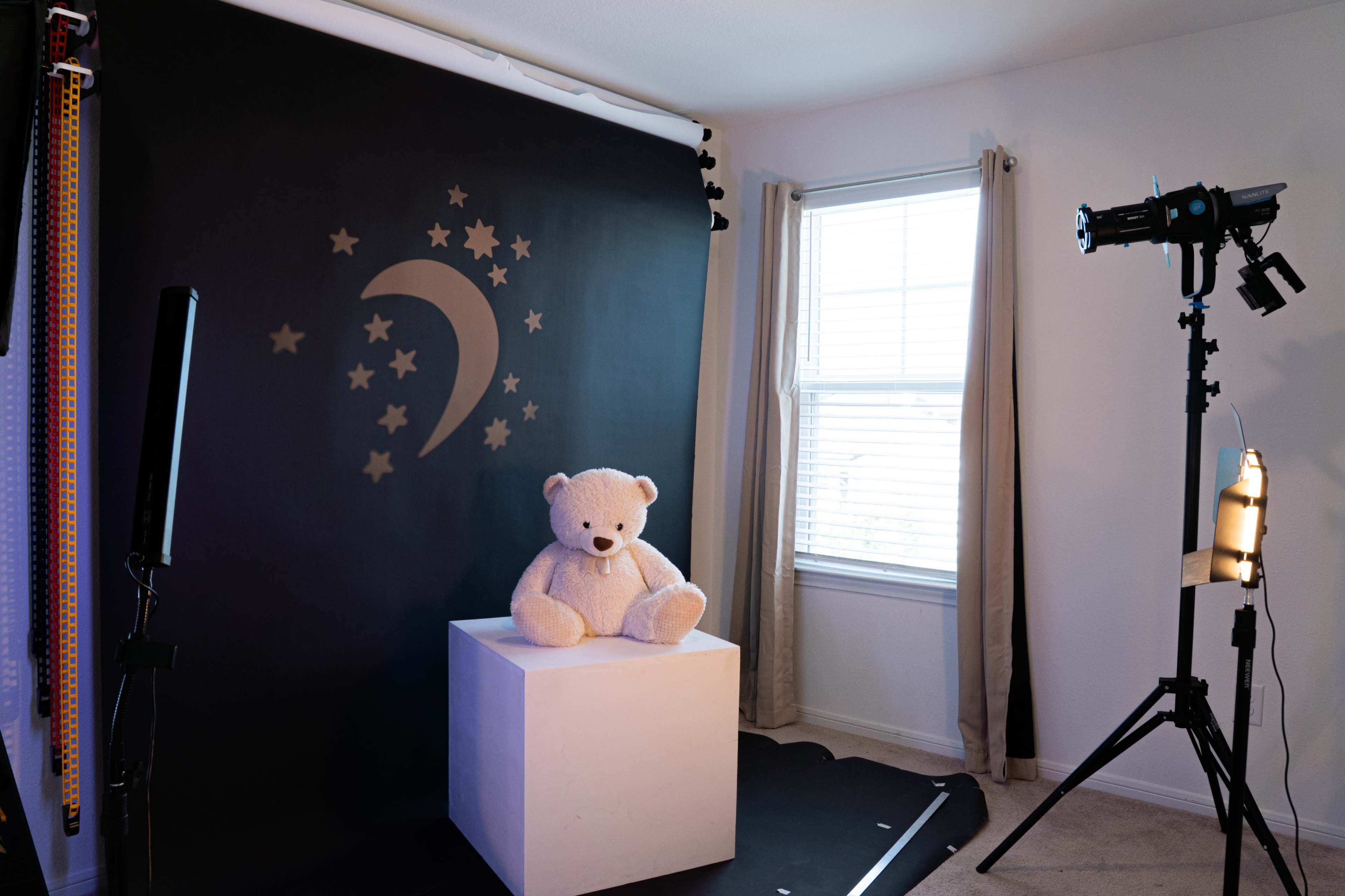 The image shows a photography studio with a plush teddy bear sitting on a white cube against a black backdrop featuring stars and a crescent moon.