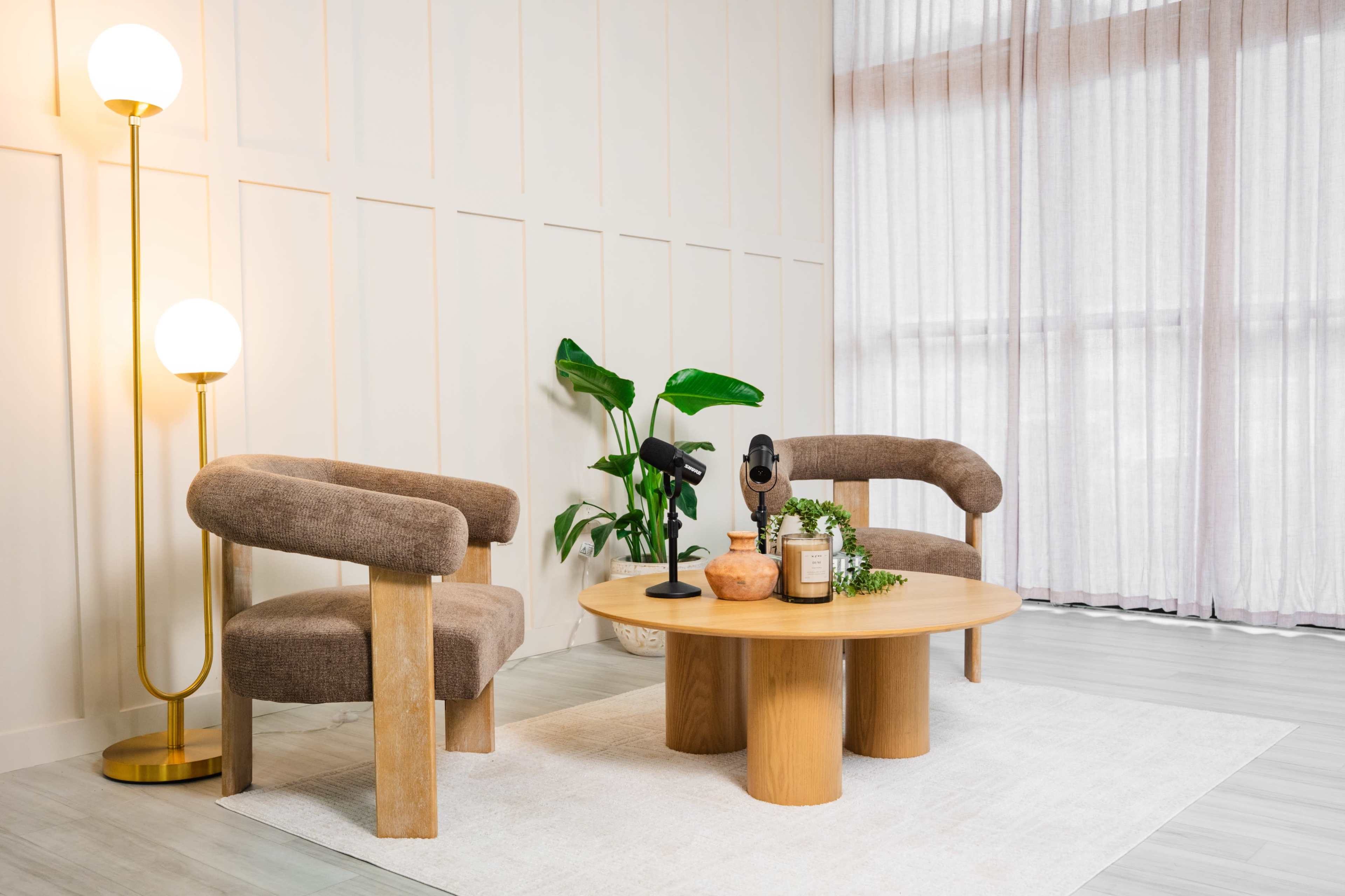 The image showcases a modern living room setup featuring two rounded chairs, a circular wooden coffee table, and a decorative plant near a tall lamp and large windows with sheer curtains.