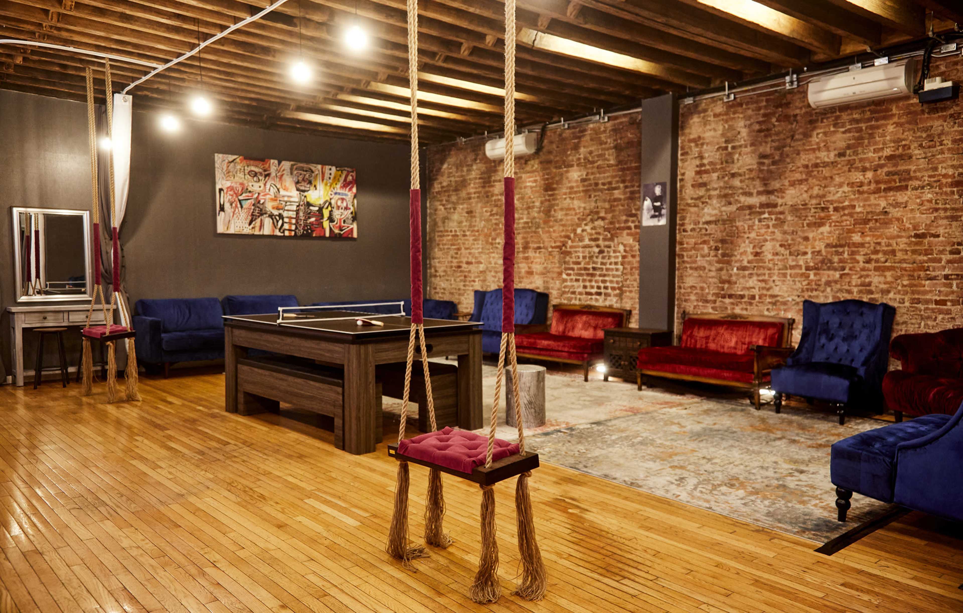 A spacious lounge area featuring a pool table, vintage-style seating in red and blue, a large rug, and swings hanging from the ceiling, all set against a brick wall.