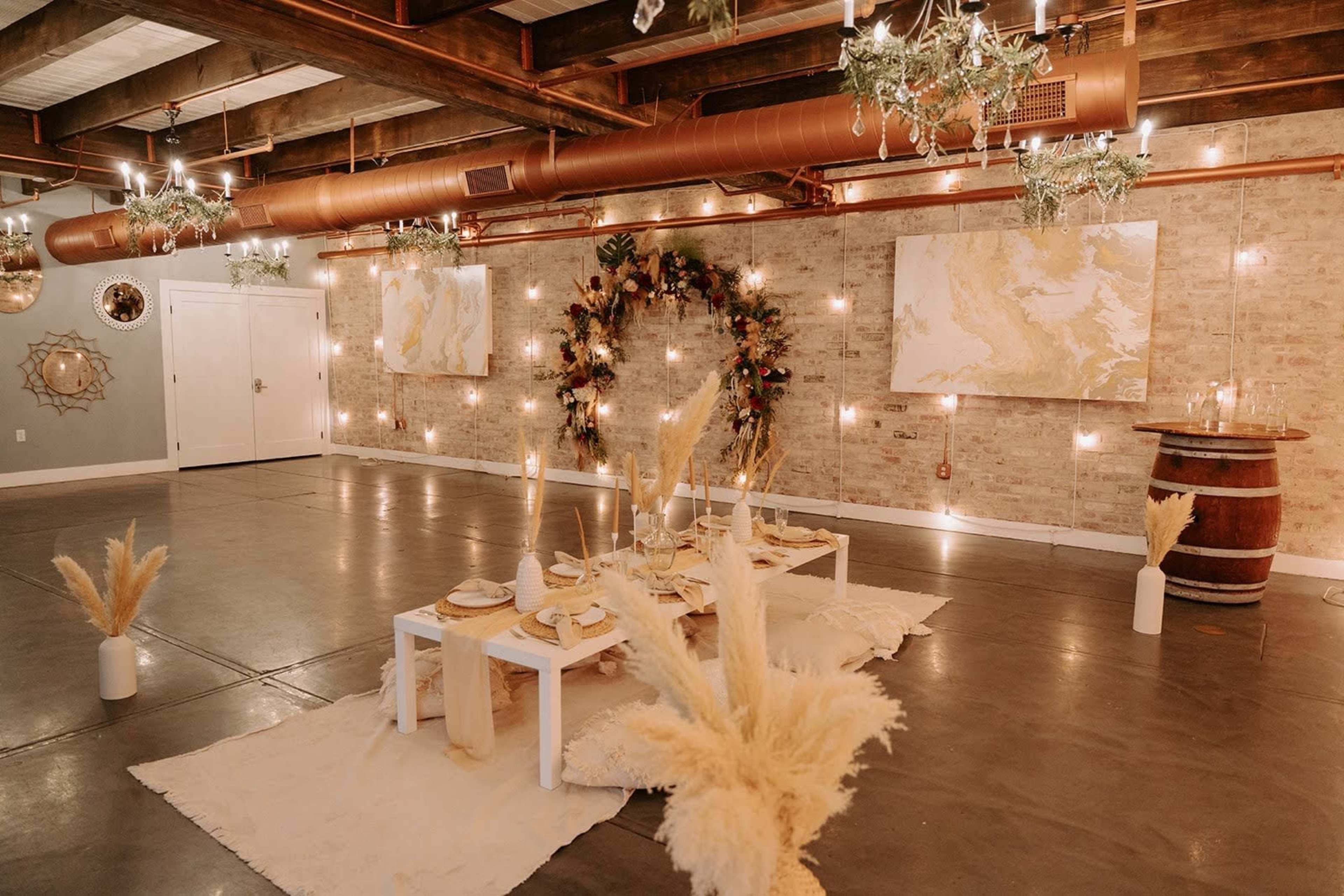 A spacious event venue features a low dining setup with a white table, decorated with gold-accented dishware, surrounded by dried flowers and soft lighting on exposed brick walls.