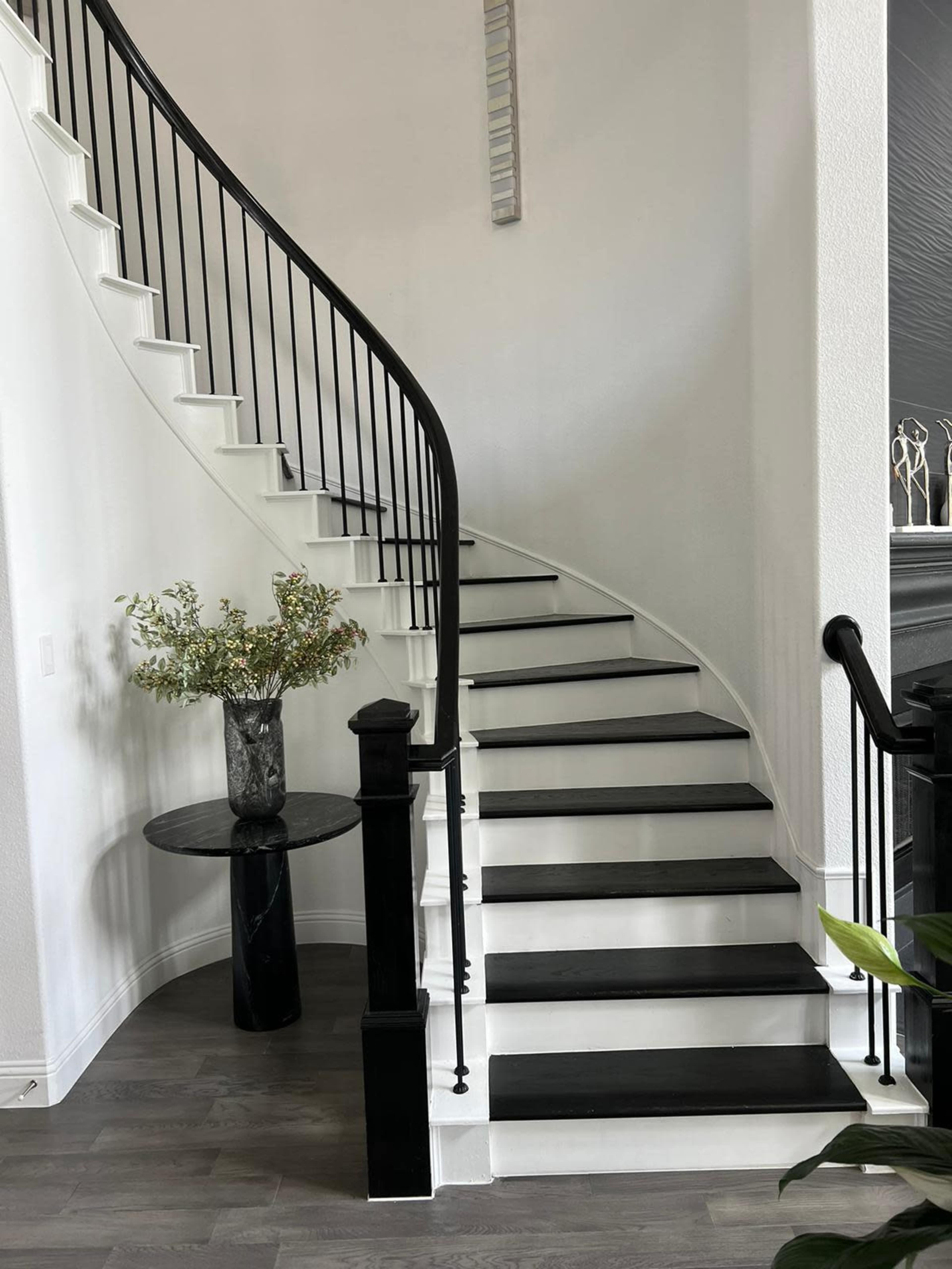 A curved staircase with black and white steps is positioned beside a small black table adorned with a floral arrangement in a well-lit entryway.