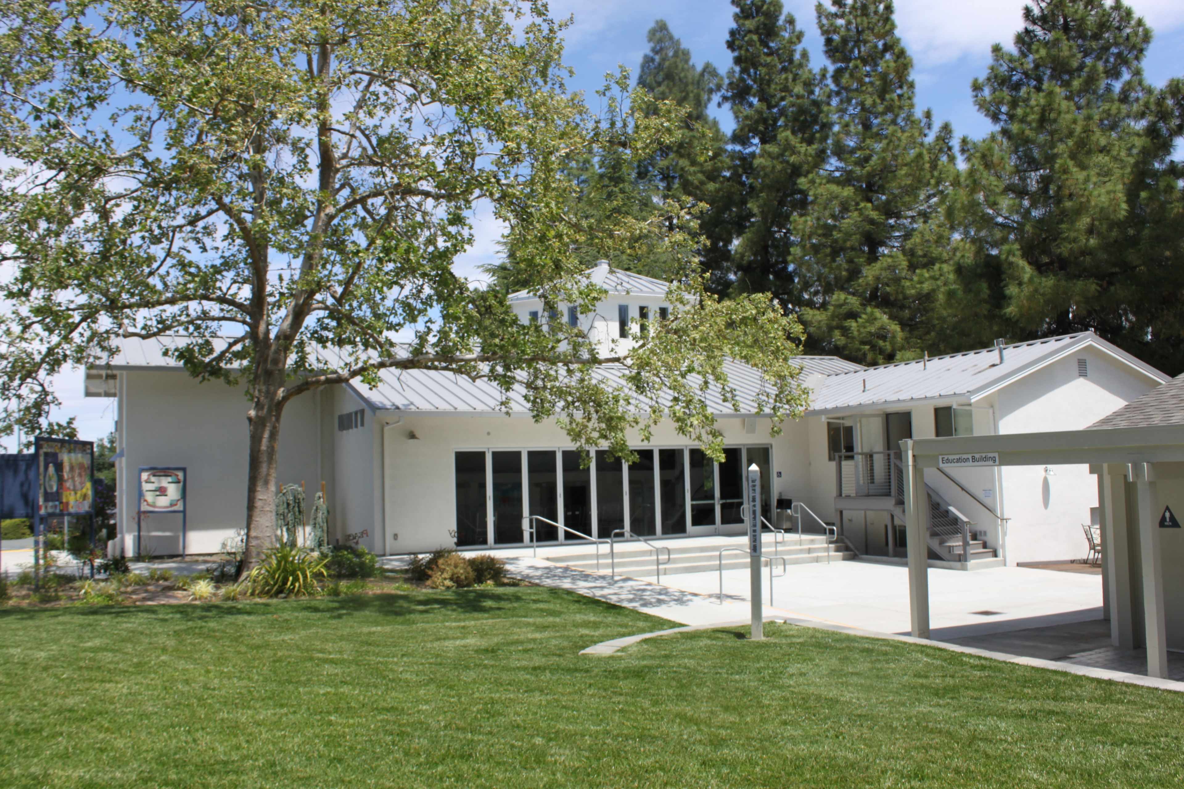 A white building with large windows and a ramp, surrounded by grass and trees on a sunny day.