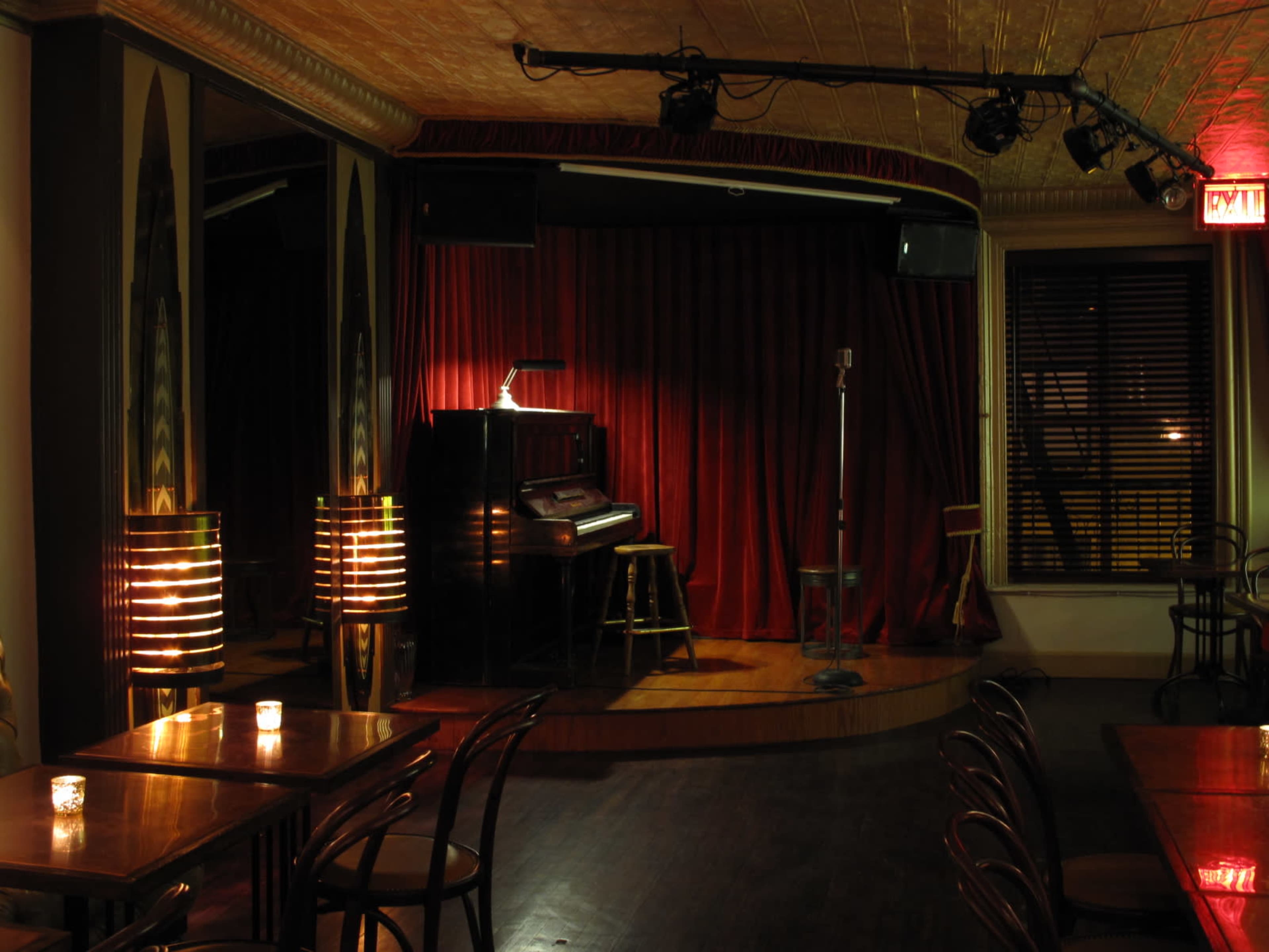 The image depicts a dimly lit performance stage in a small venue, featuring a red curtain, a vintage piano, and a microphone on a stand.