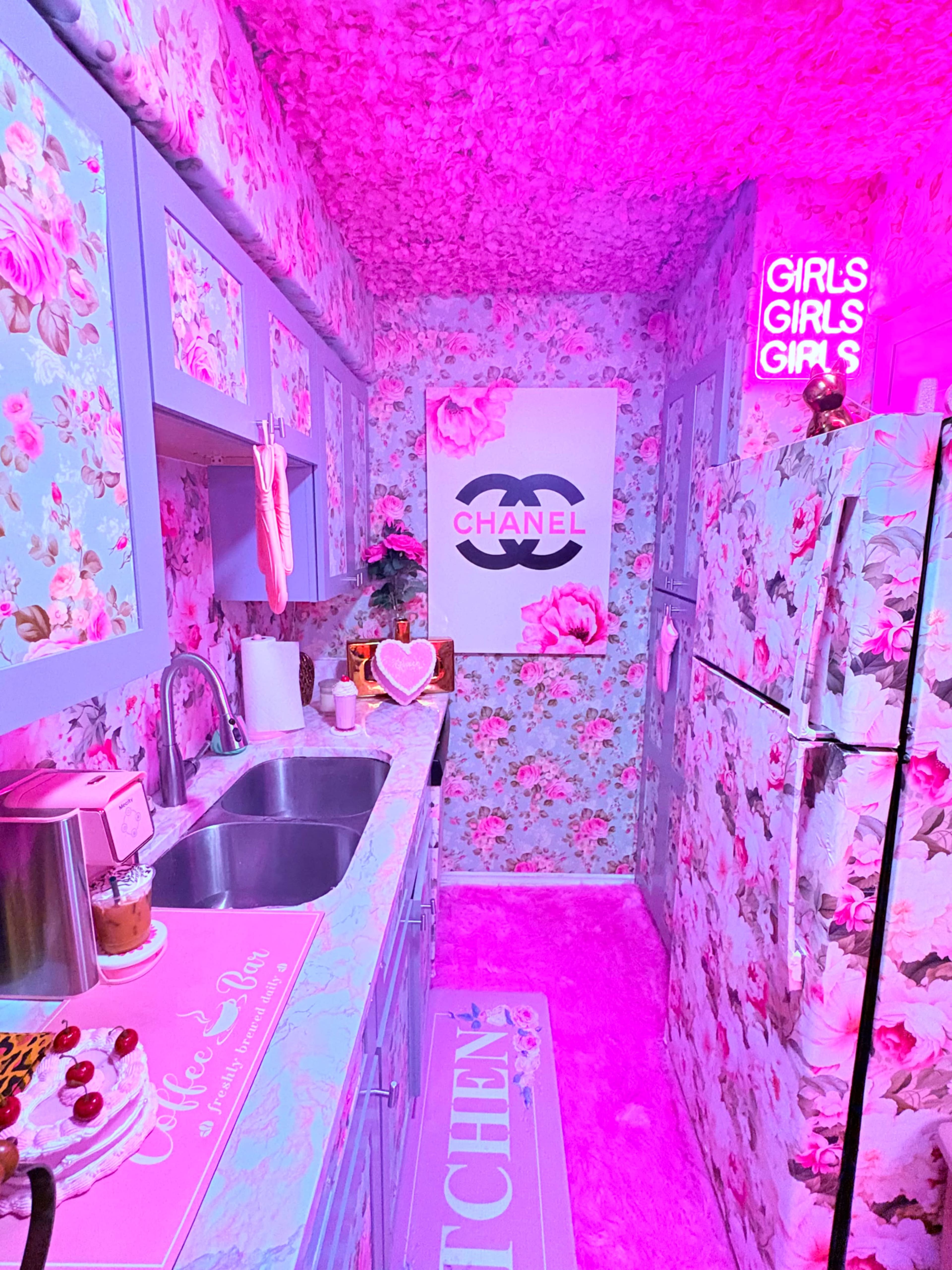 The image shows a small, brightly colored kitchen with floral wallpaper, pink lighting, and a Chanel-themed art piece on the wall.