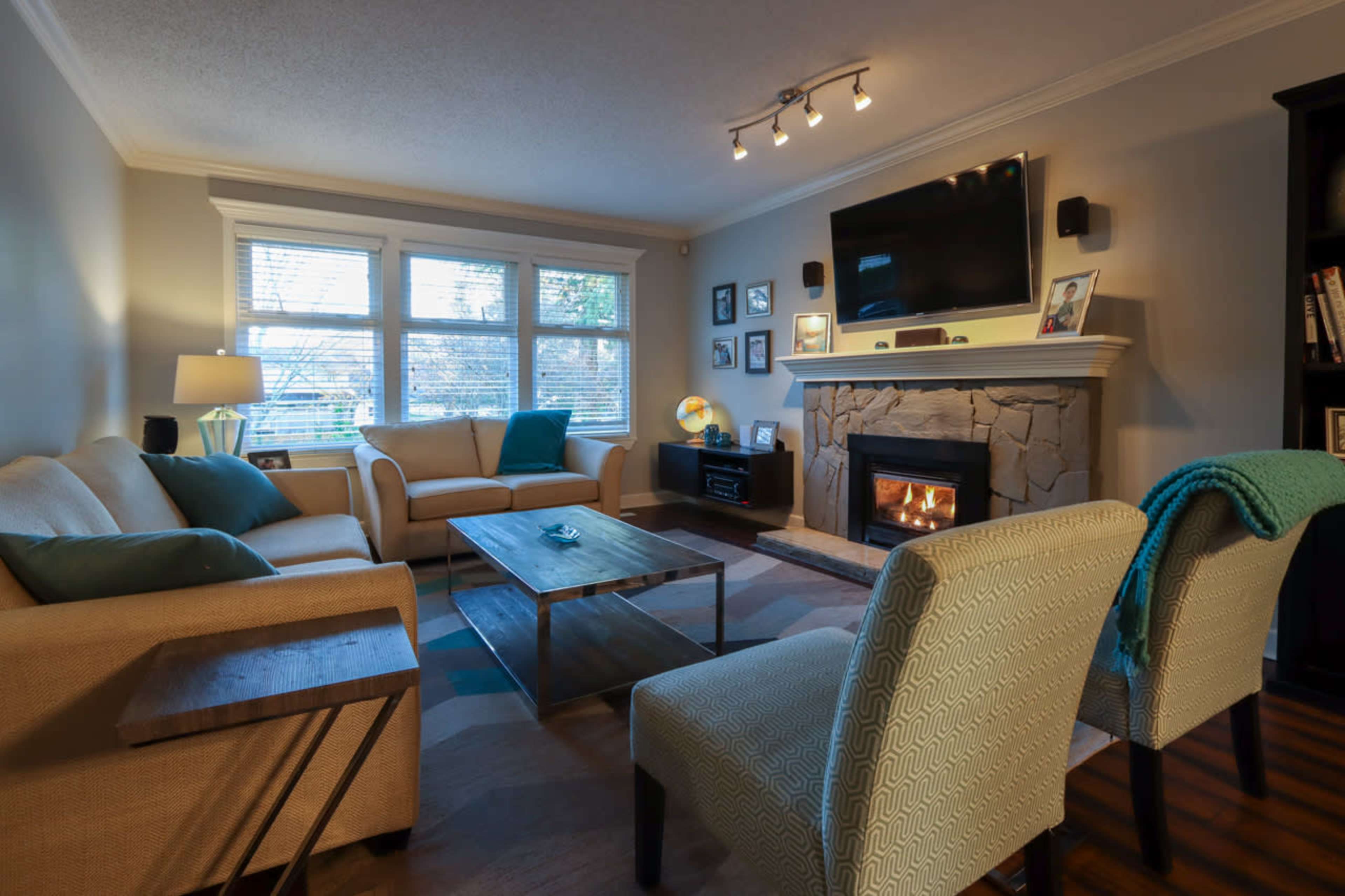 A living room features a beige sofa set, a stone fireplace with a TV mounted above, and a wooden coffee table, illuminated by natural light from the windows.