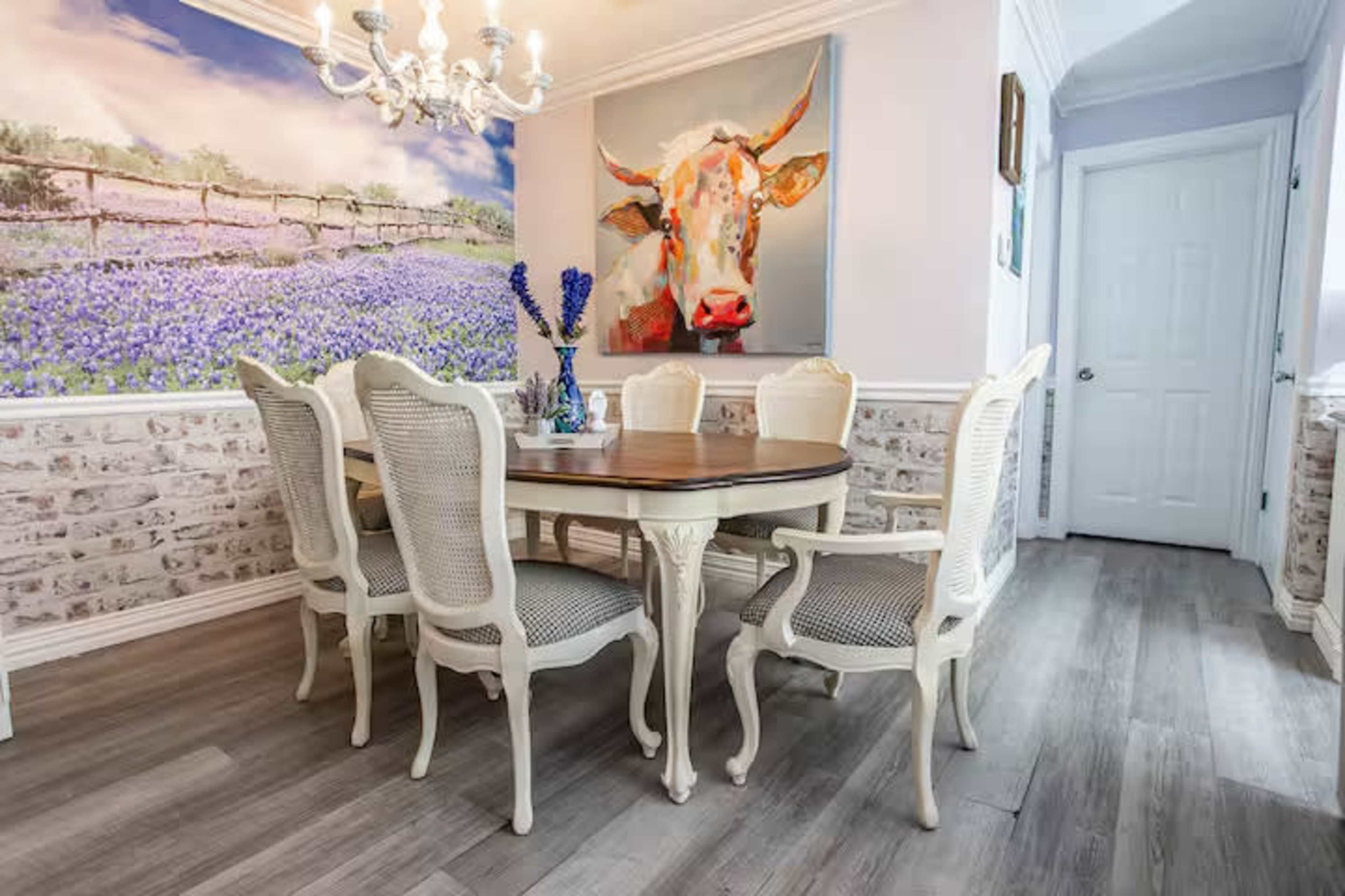 The image shows a dining area with a large wooden table surrounded by white, classical chairs, a chandelier above, and a colorful cow painting on the wall.