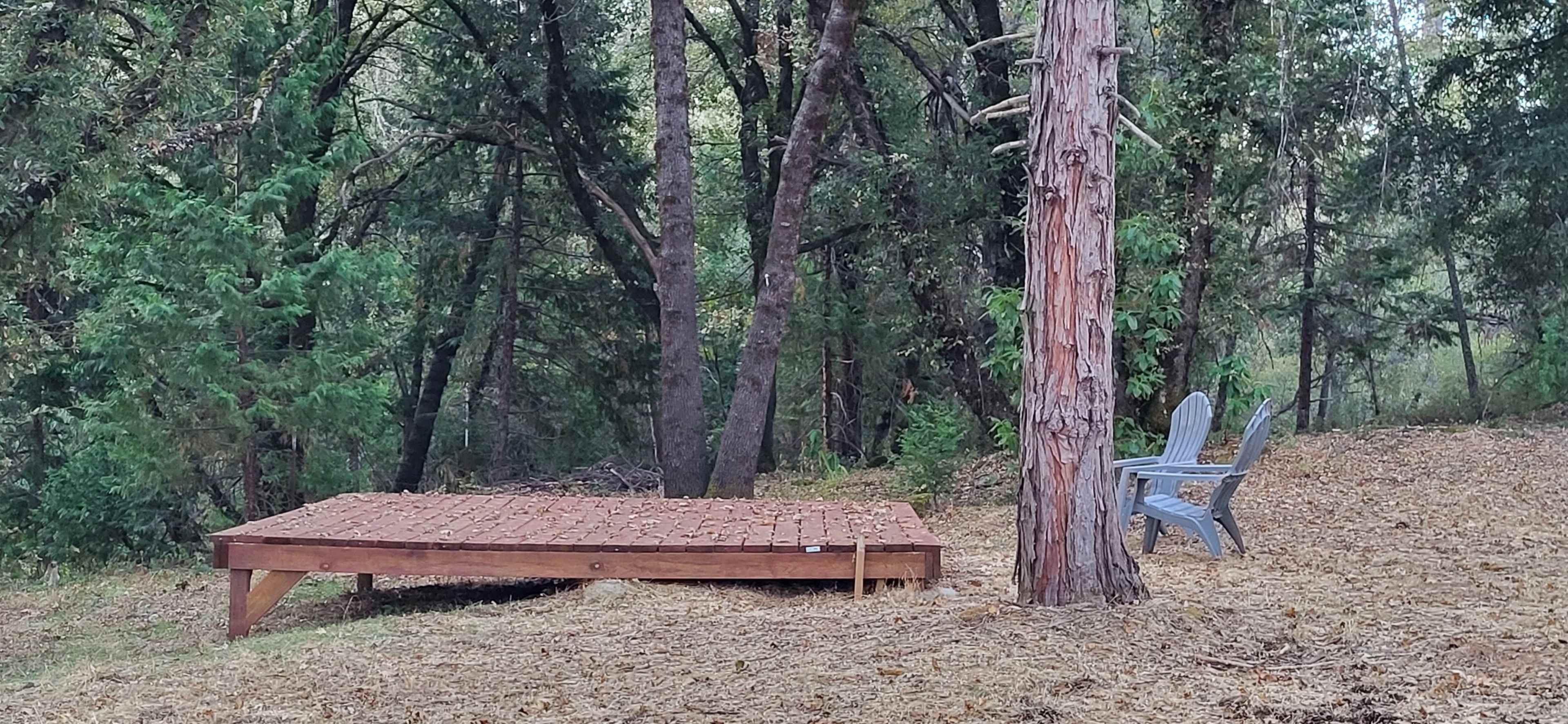 A wooden platform is situated in a forest clearing next to a single gray chair and a tall tree.