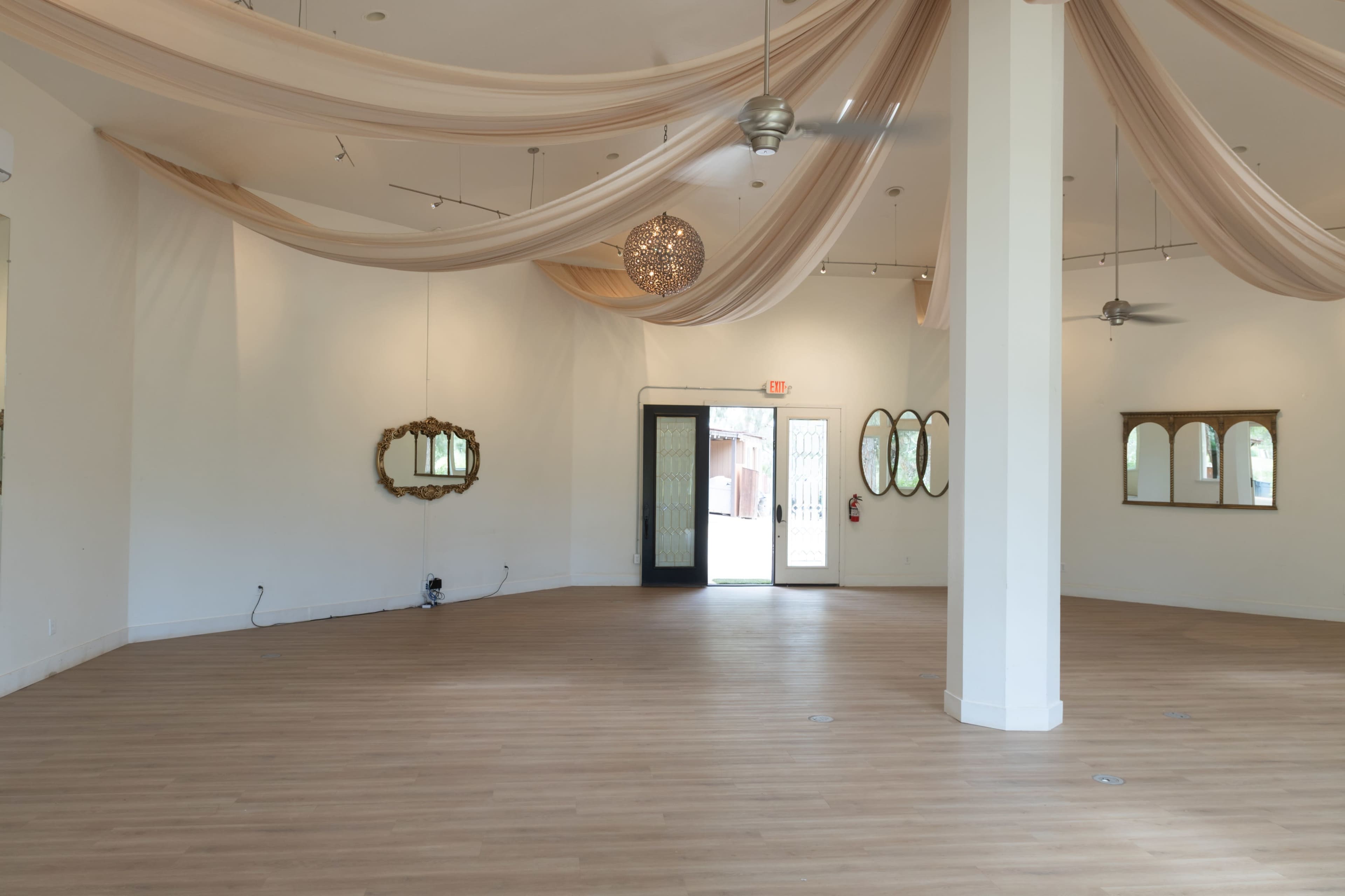 A spacious room features cascading fabric drapes from the ceiling, large mirrors on the walls, and a doorway leading outside.