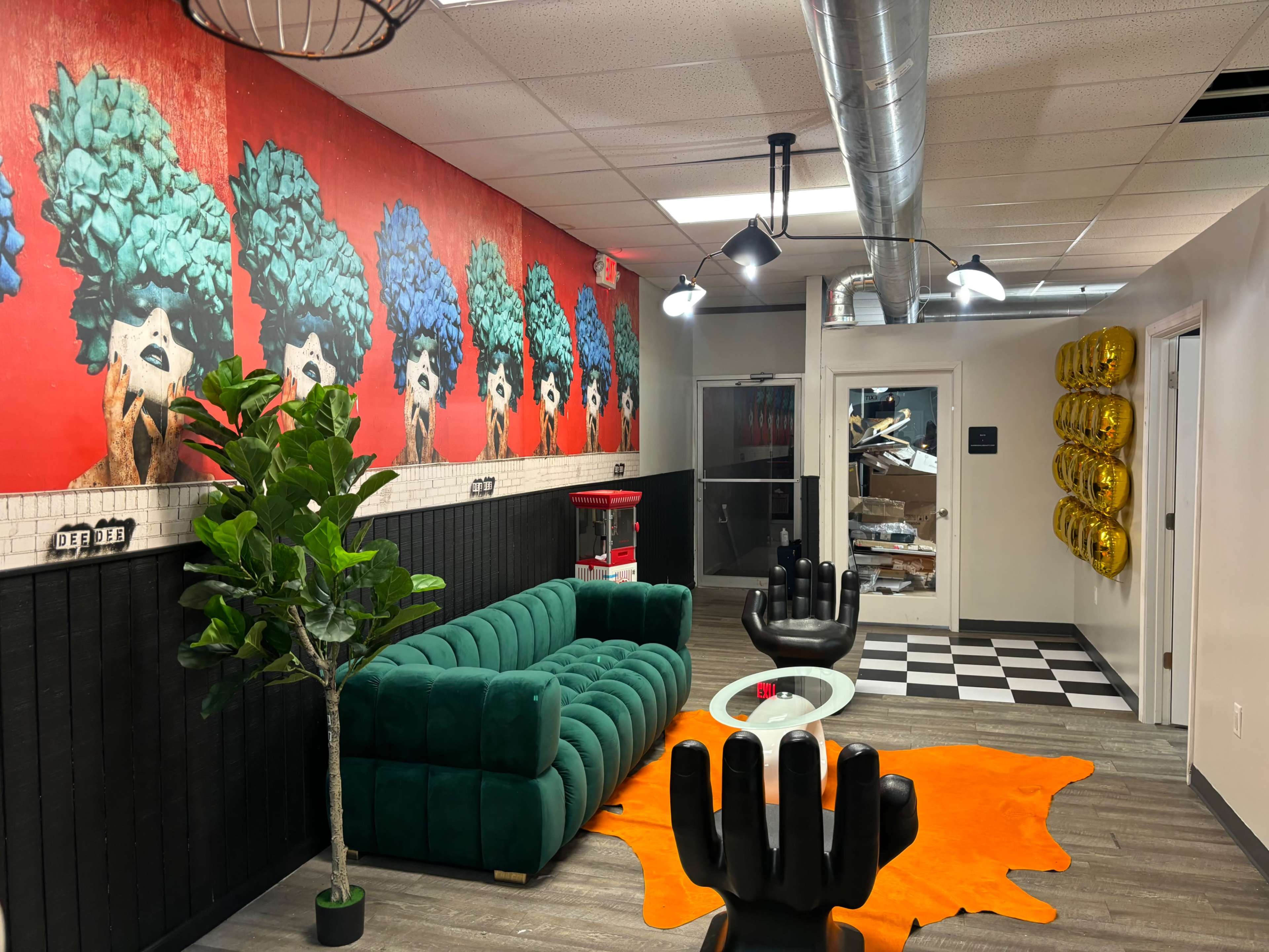 A modern salon interior featuring a green velvet sofa, black chairs, a checkered floor, and a vibrant mural of colorful abstract figures on the wall.