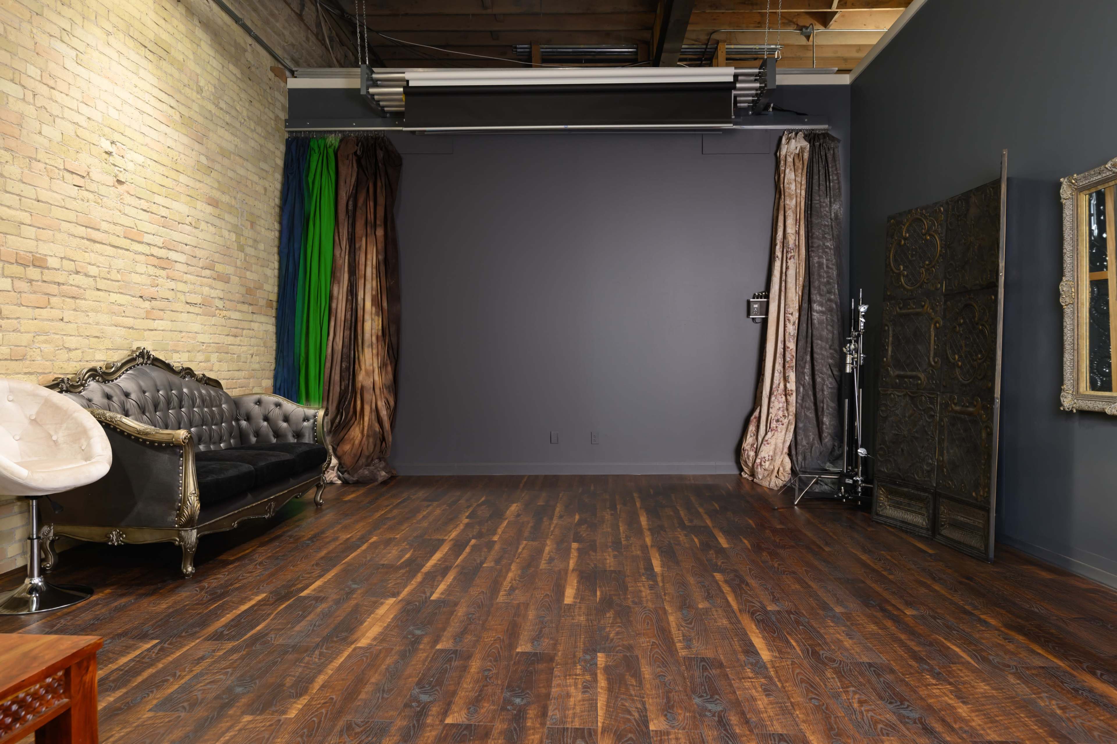 The image shows an empty studio space with hardwood flooring, a decorative couch, and various curtains on either side of a plain wall.