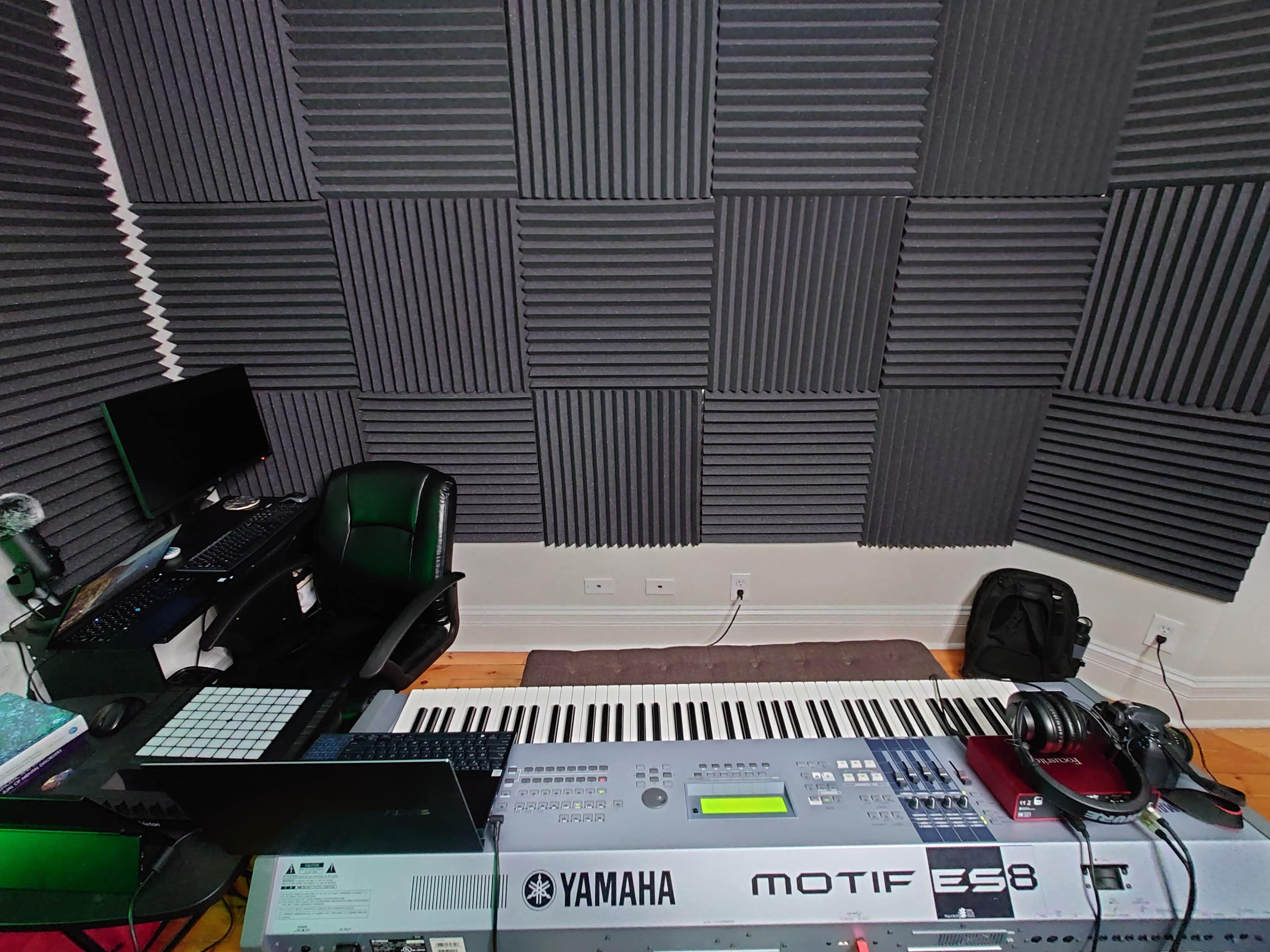 A music production setup features a Yamaha Motif ES8 keyboard, a computer, a black chair, and acoustic foam panels on the walls.
