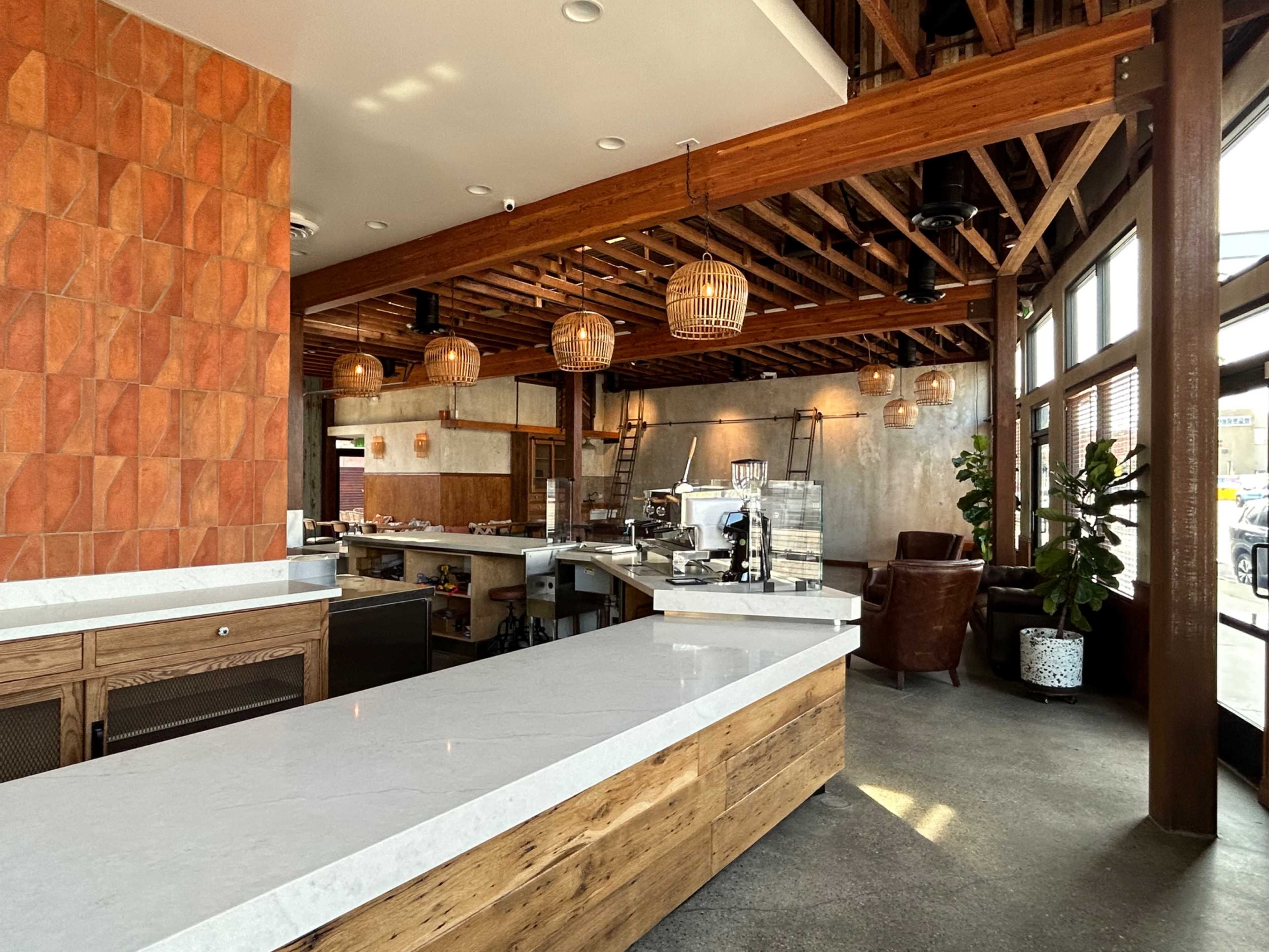 A modern cafe interior with a marble countertop, wooden accents, pendant lighting, and a seating area featuring leather chairs and potted plants.
