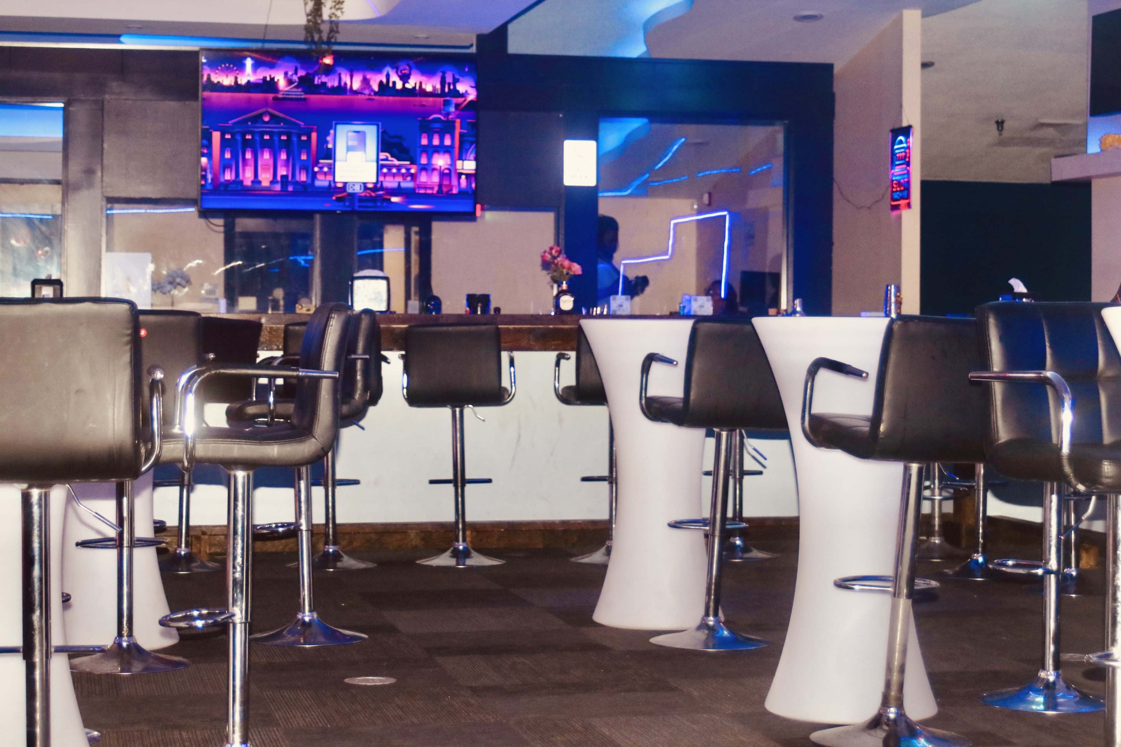 The scene shows a modern bar area with white high-top tables and black chairs, illuminated by blue lighting, and featuring a television mounted on the wall displaying images.