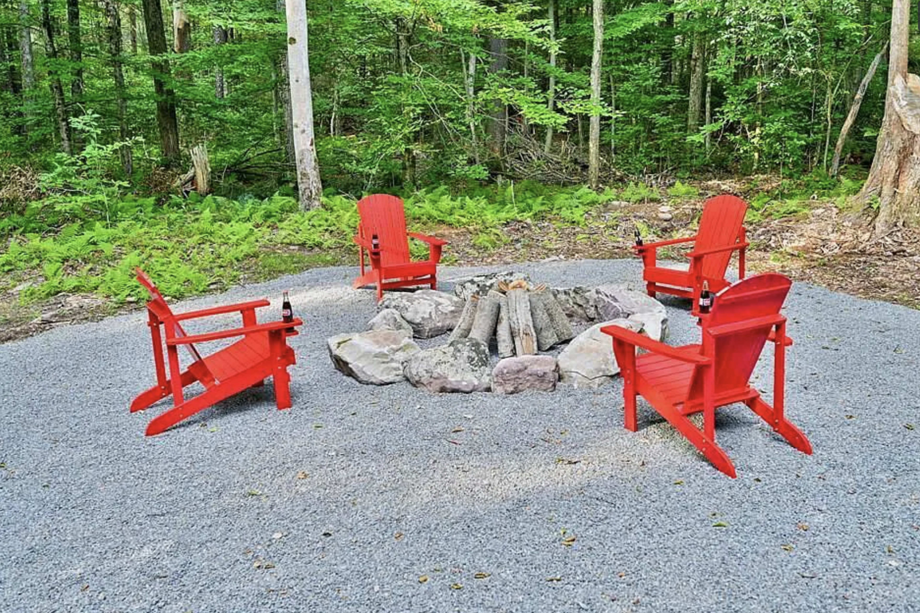 A circular arrangement of four red Adirondack chairs is set around a stone fire pit in a wooded area.