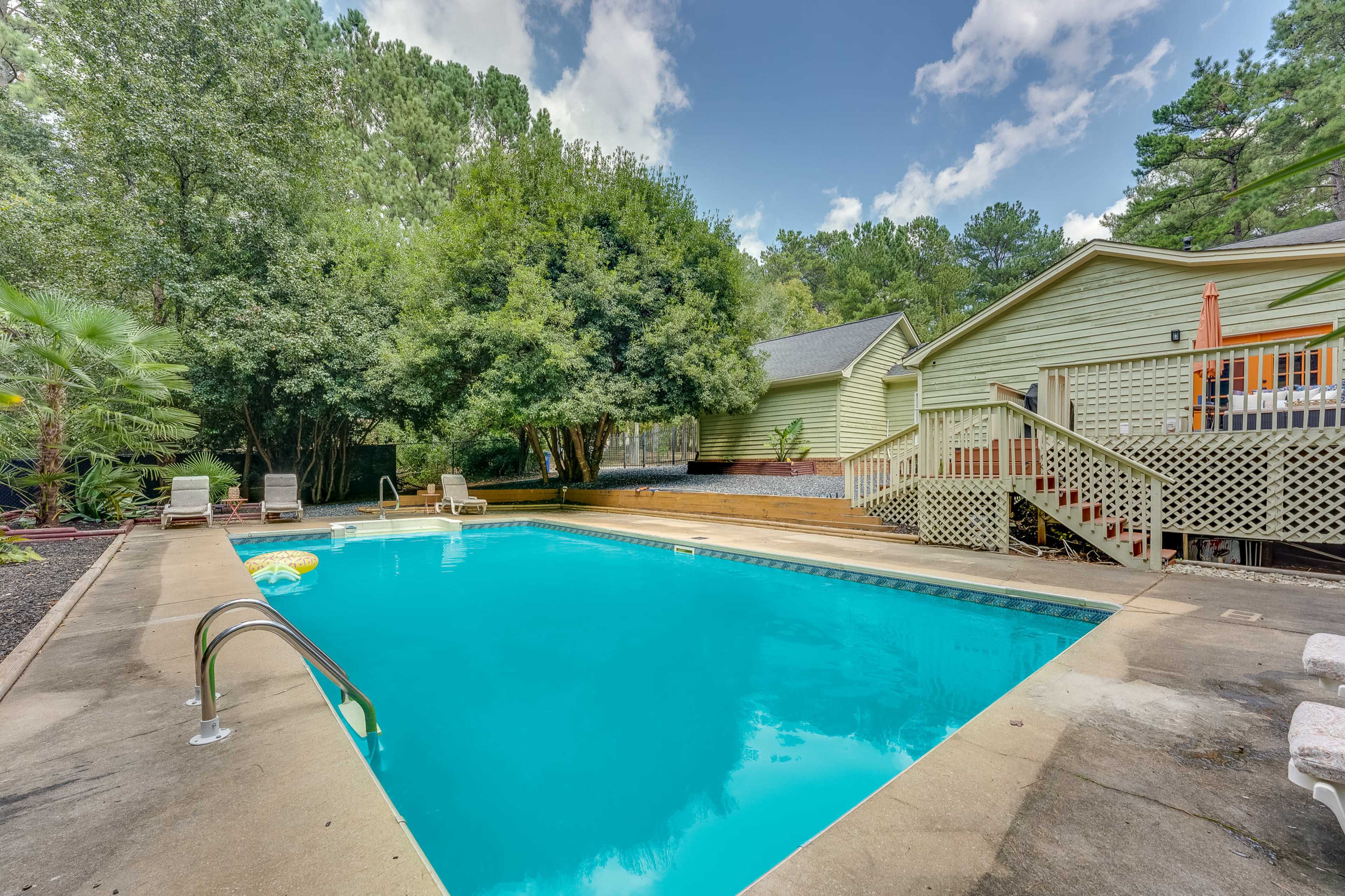 The image shows a clear swimming pool surrounded by concrete, with lounge chairs nearby and a house partially visible in the background.