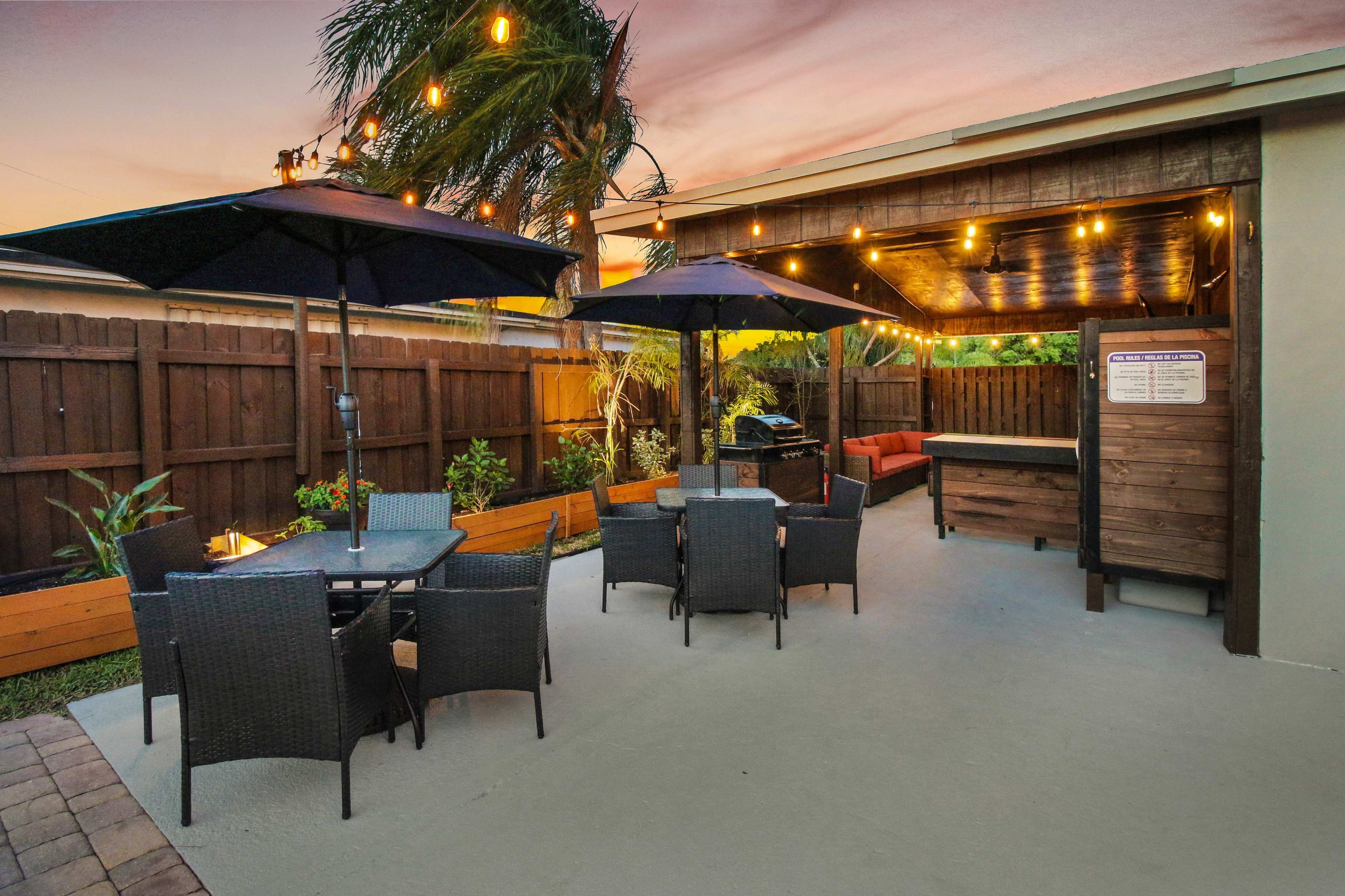 The image shows a backyard patio area with outdoor furniture, umbrellas, and string lights overhead during sunset.