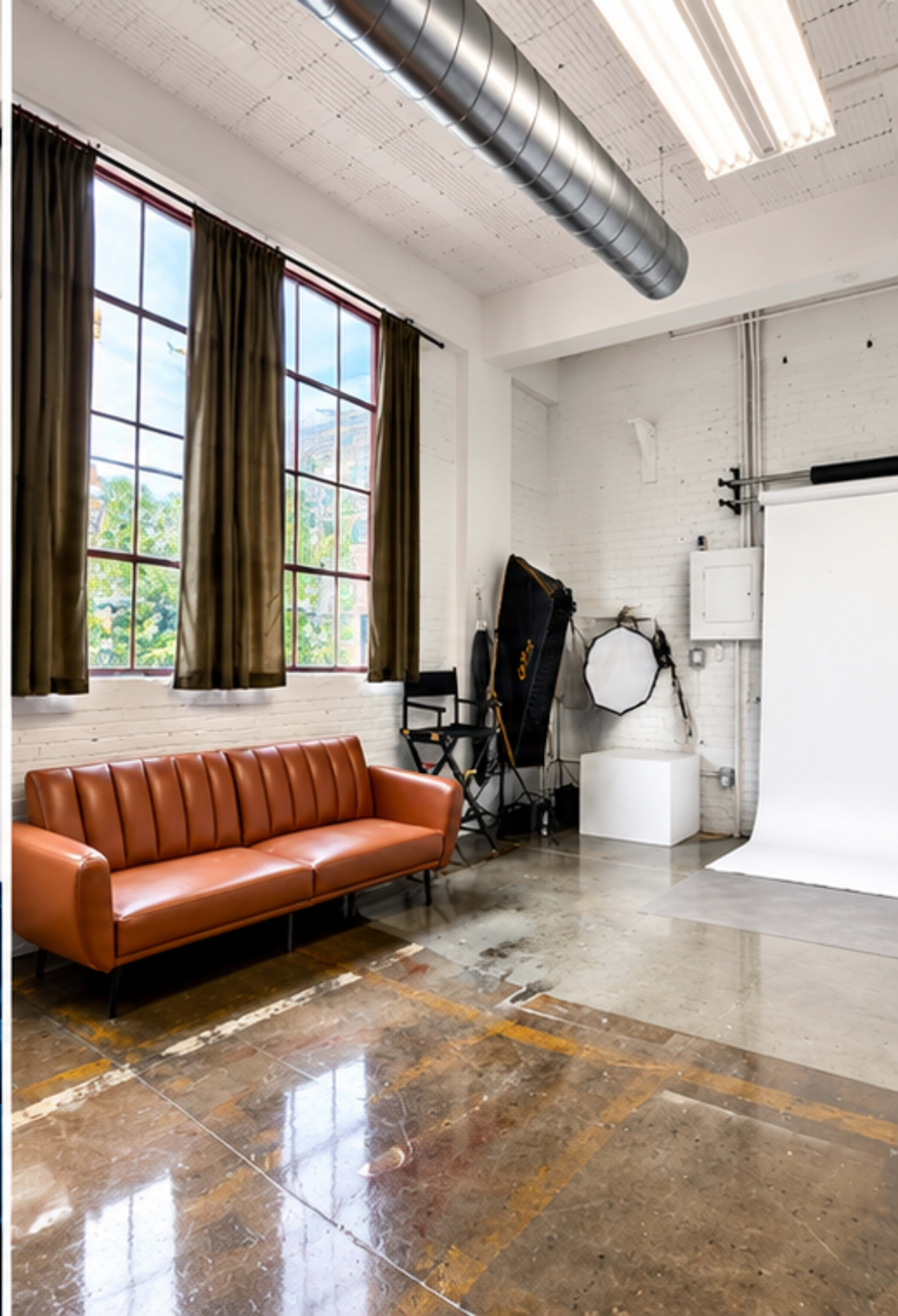 The image shows a spacious photography studio with a brown leather sofa, large windows, and a backdrop setup against a white wall.