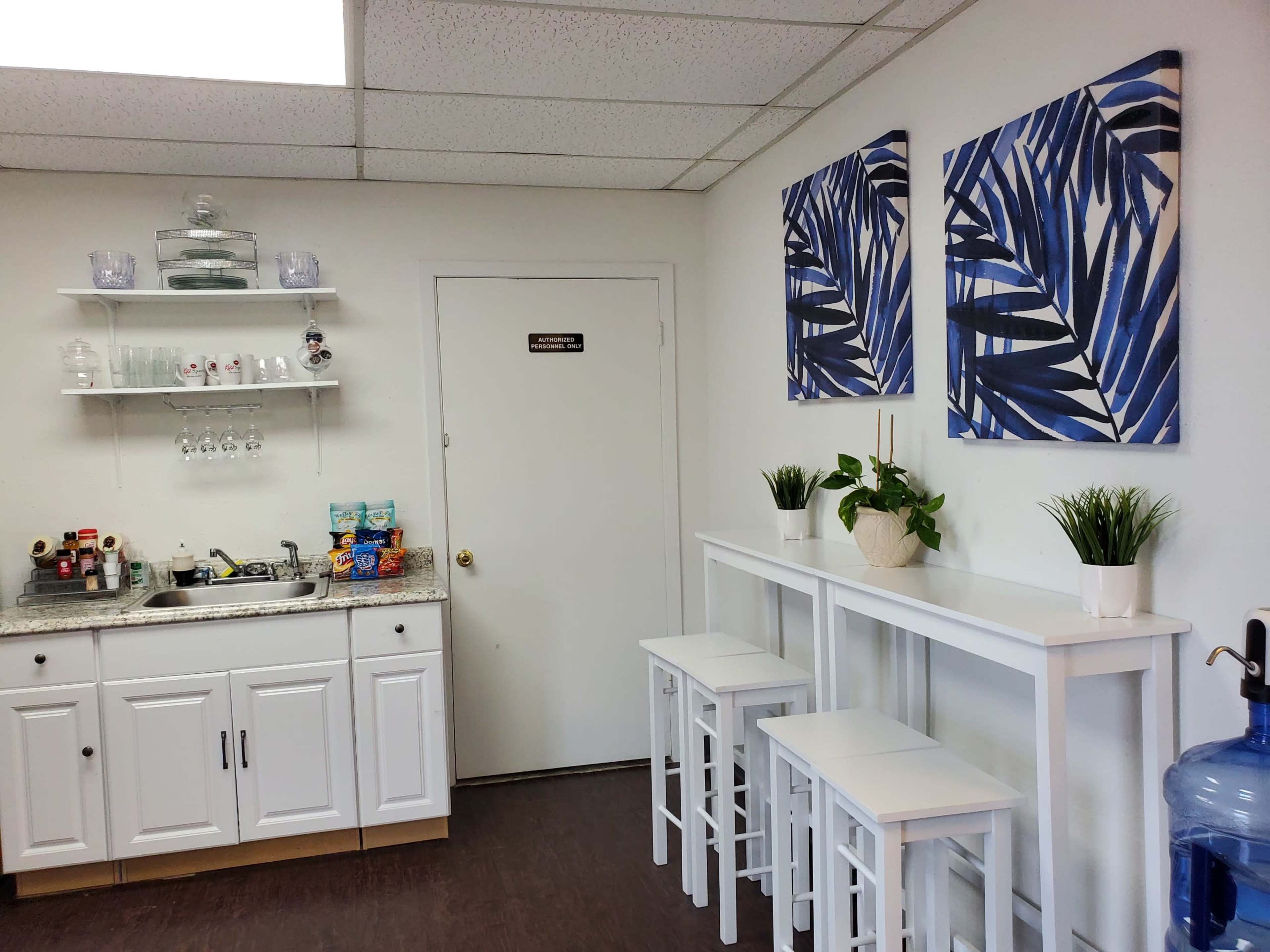 The image shows a kitchenette area with a countertop, a sink, two paintings of blue leaves on the wall, a small table with three stools, and a water dispenser.