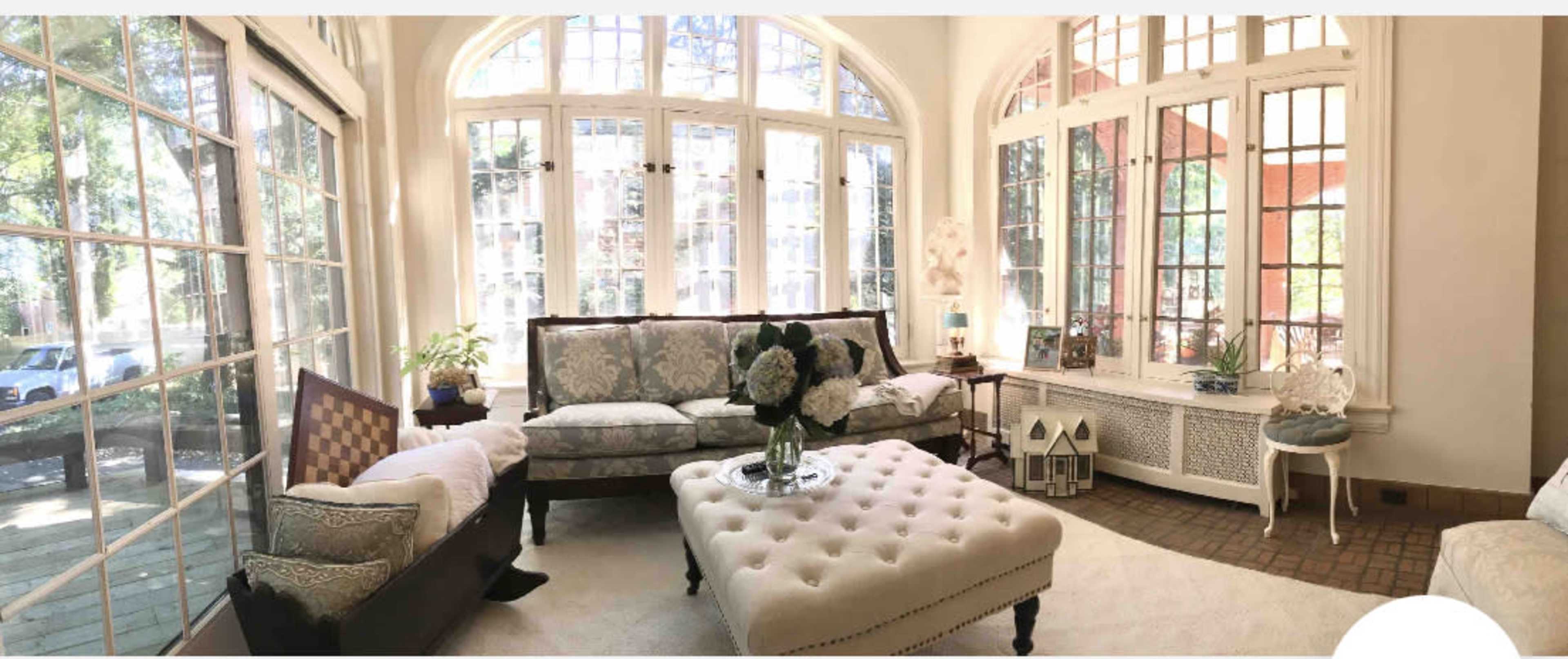 A sunlit living room with large windows, a plush beige ottoman, and elegant seating arranged around a coffee table.