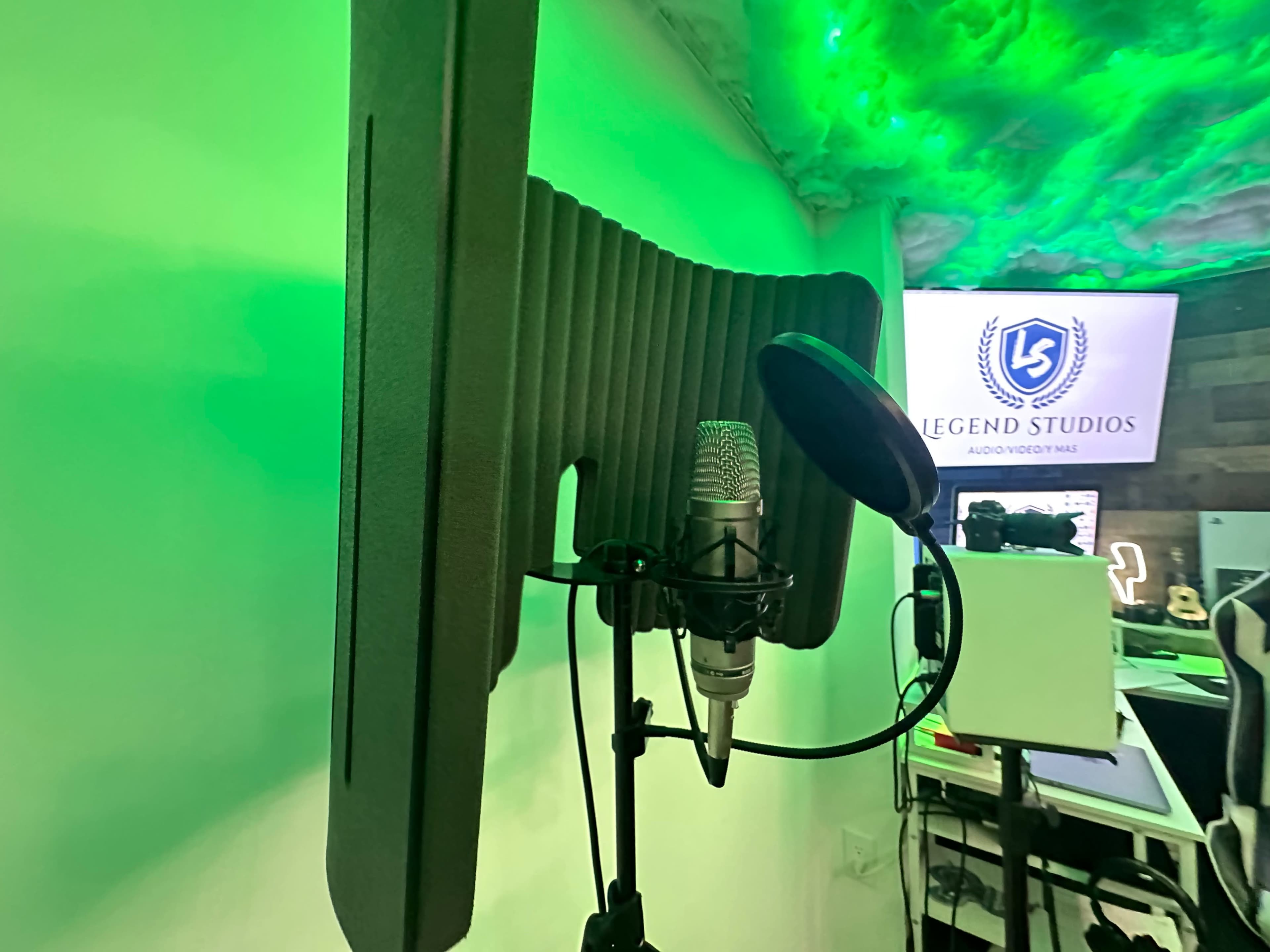 A microphone setup with a pop filter in front of a sound-absorbing panel, illuminated by green lighting, with a sign for "Legend Studios" in the background.