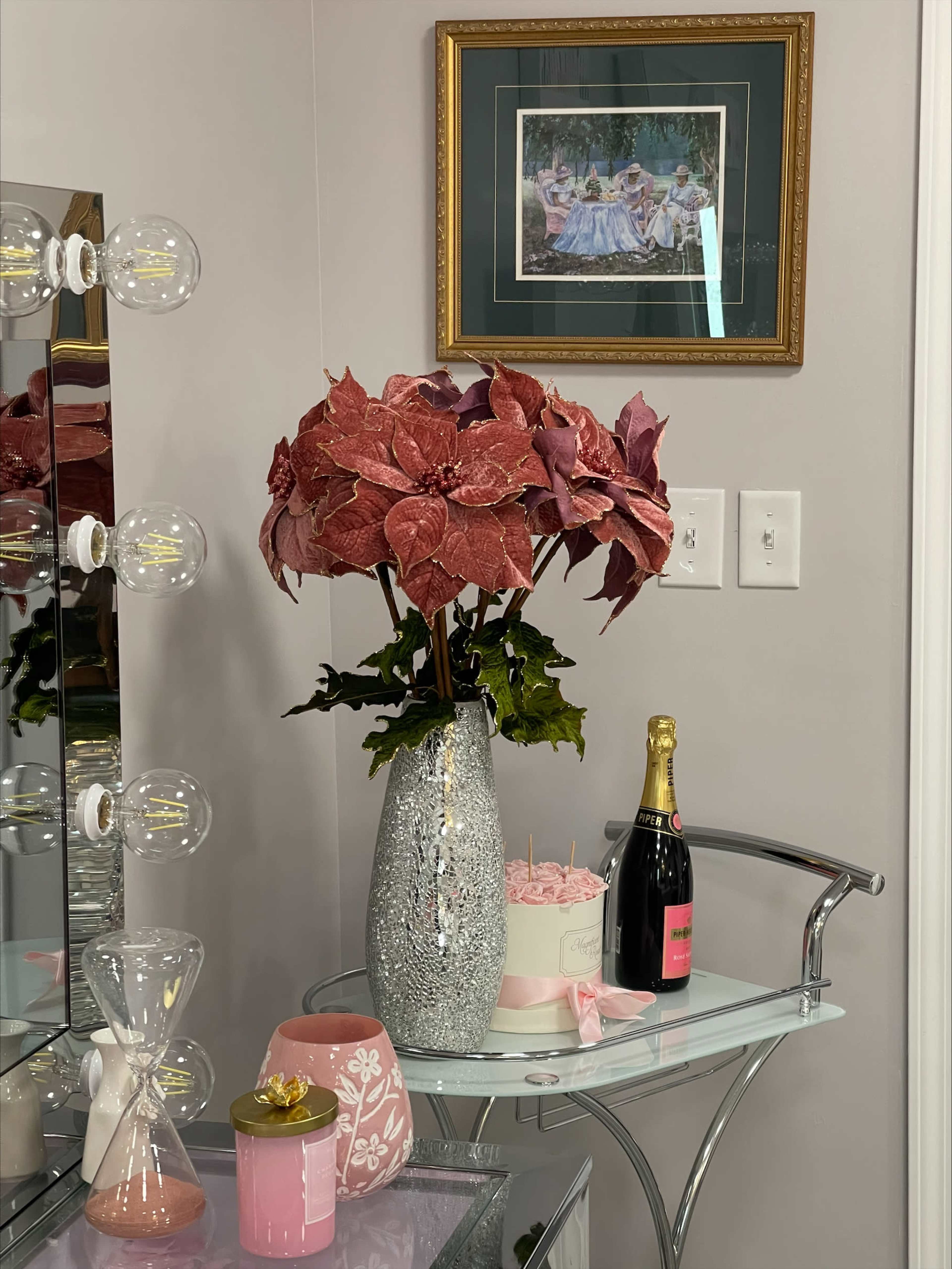A decorative vanity features a silver vase with pink foliage, a bottle of champagne, and a small cake on a glass table, with a framed artwork hanging on the wall above.