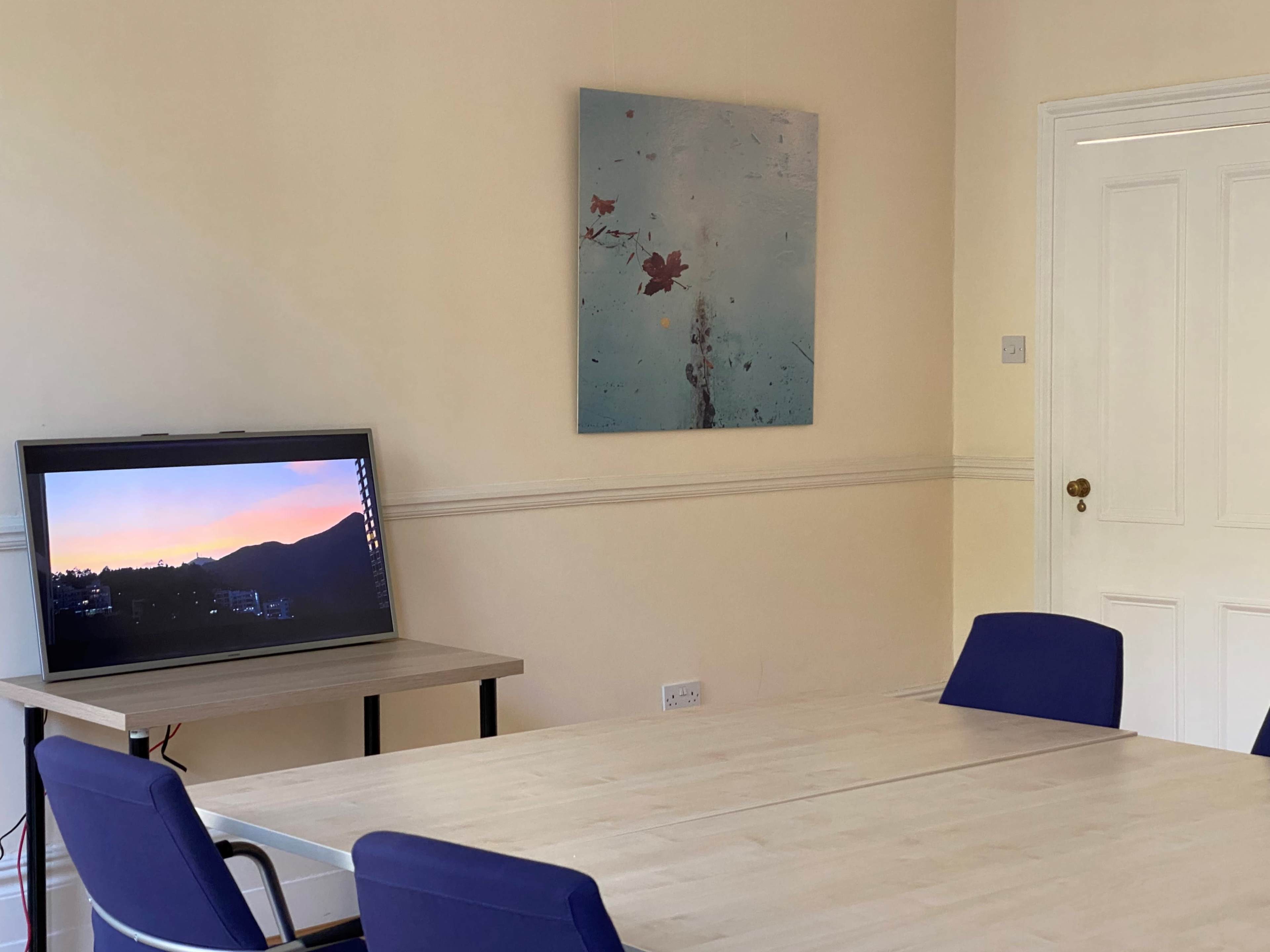 A wooden table with four blue chairs is set in a room featuring a television displaying a sunset and a wall-mounted painting.