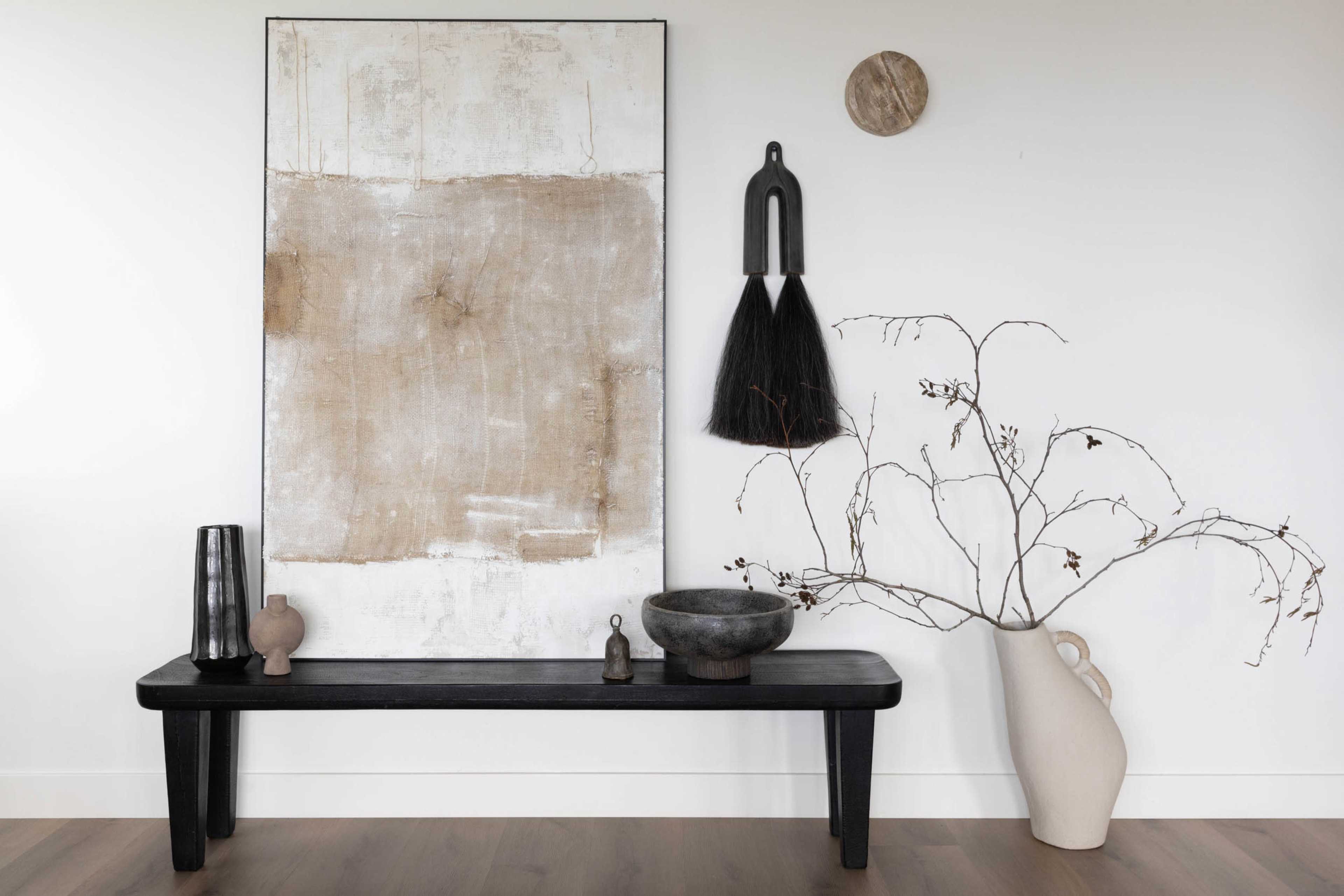 The image features a minimalist interior scene with a black wooden bench displaying various decorative objects, including vases and a bowl, alongside a large abstract artwork on the wall.
