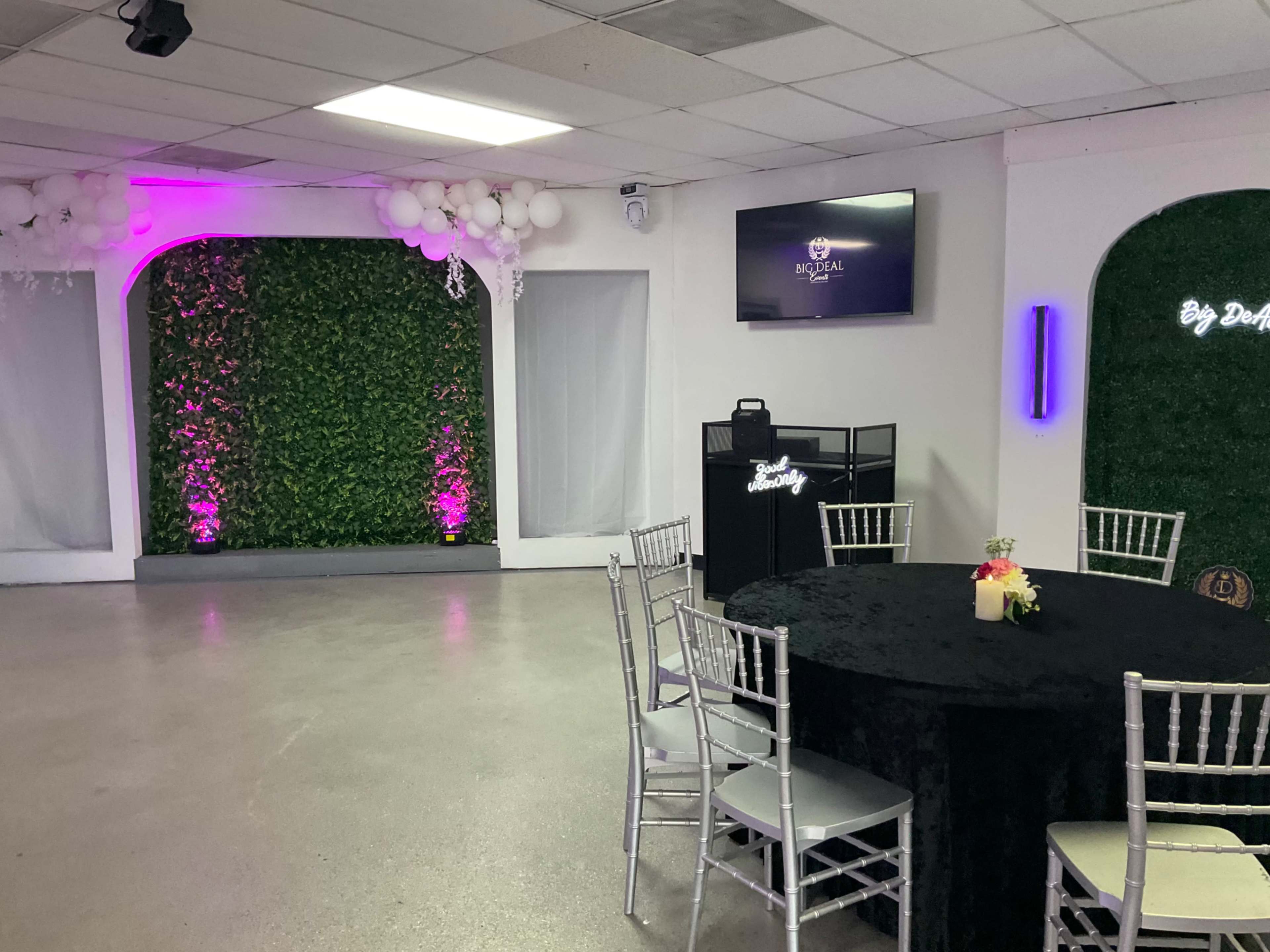 The image shows a banquet room featuring a round black table with silver chairs, a green wall with decorative foliage, and ambient purple lighting.