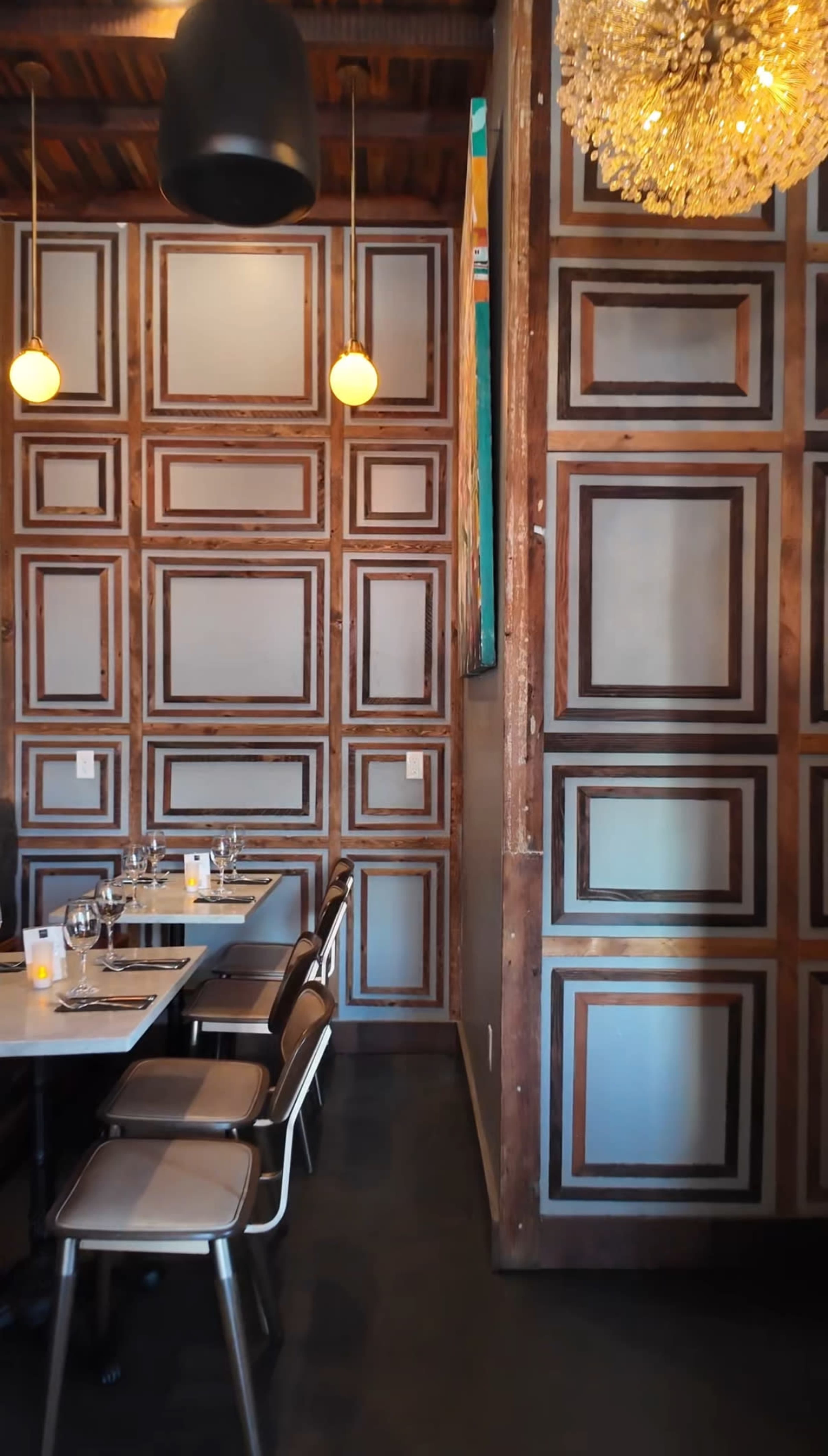 The image depicts a restaurant interior with a wall featuring a geometric pattern of framed panels and a dining table set with glasses and cutlery.