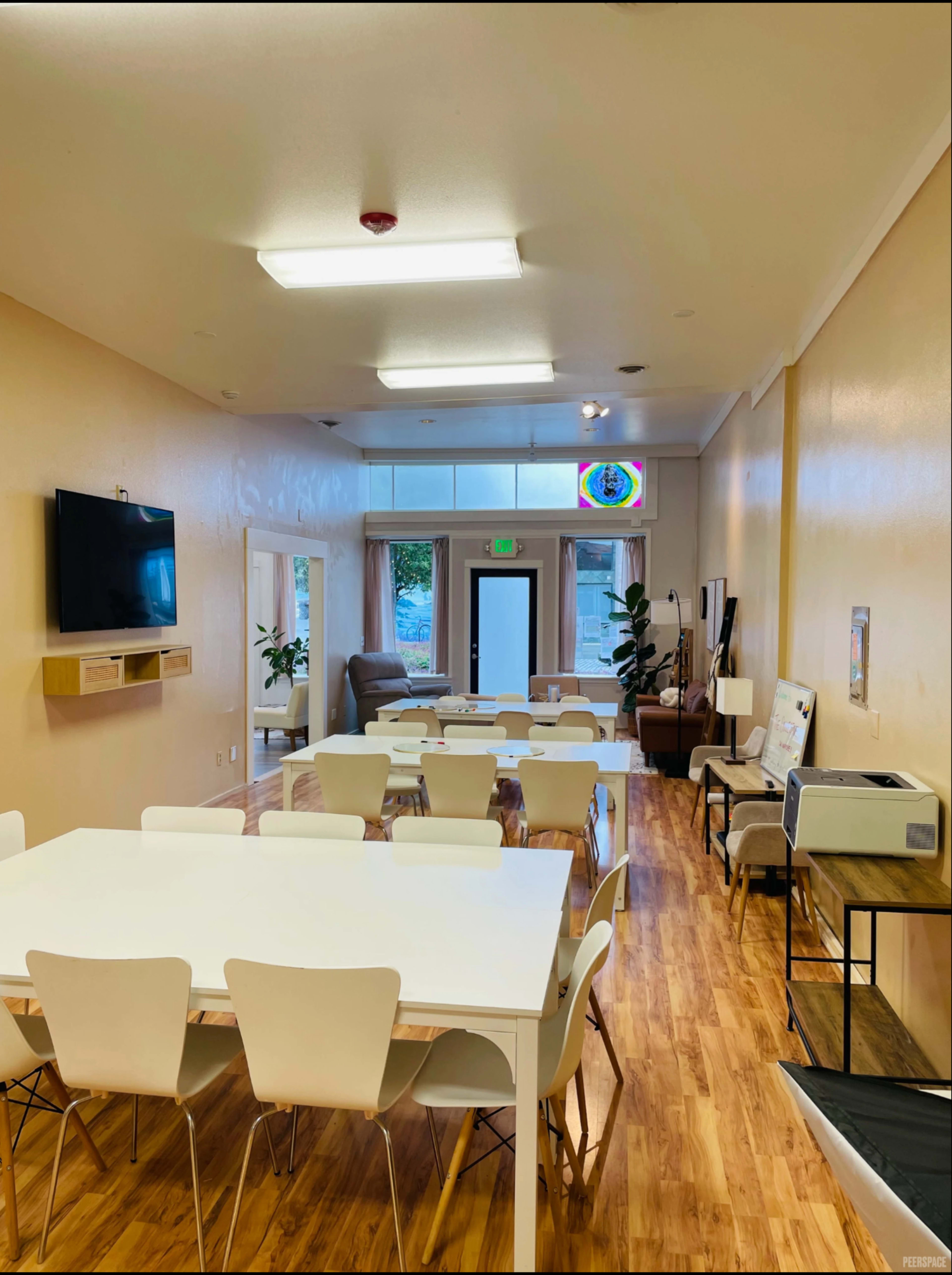 The image shows a spacious, well-lit interior of a communal room featuring a large dining table, a seating area, and a television on the wall.