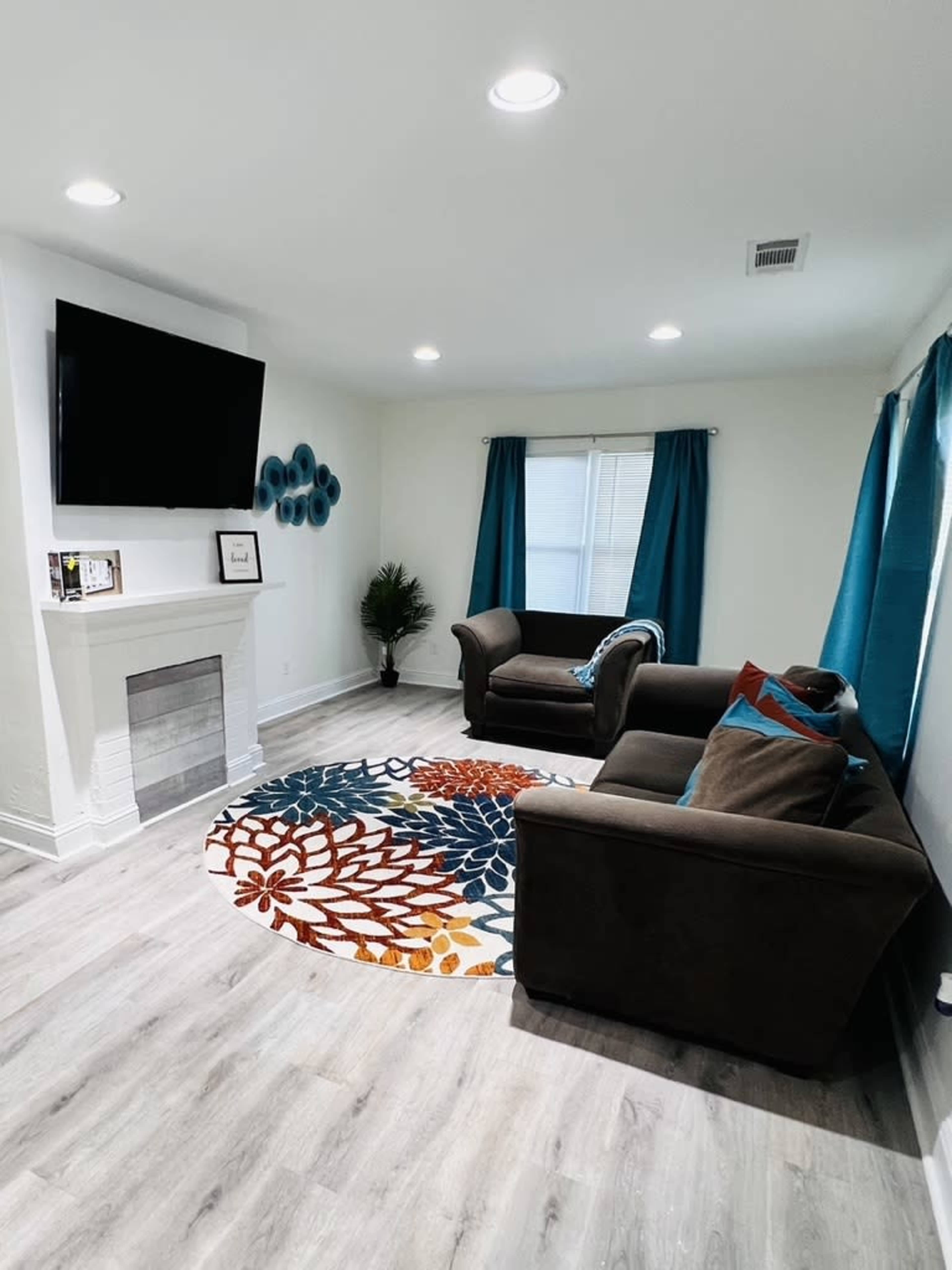 A modern living room with two brown sofas, a colorful circular rug, a television mounted on the wall, and blue curtains covering the windows.