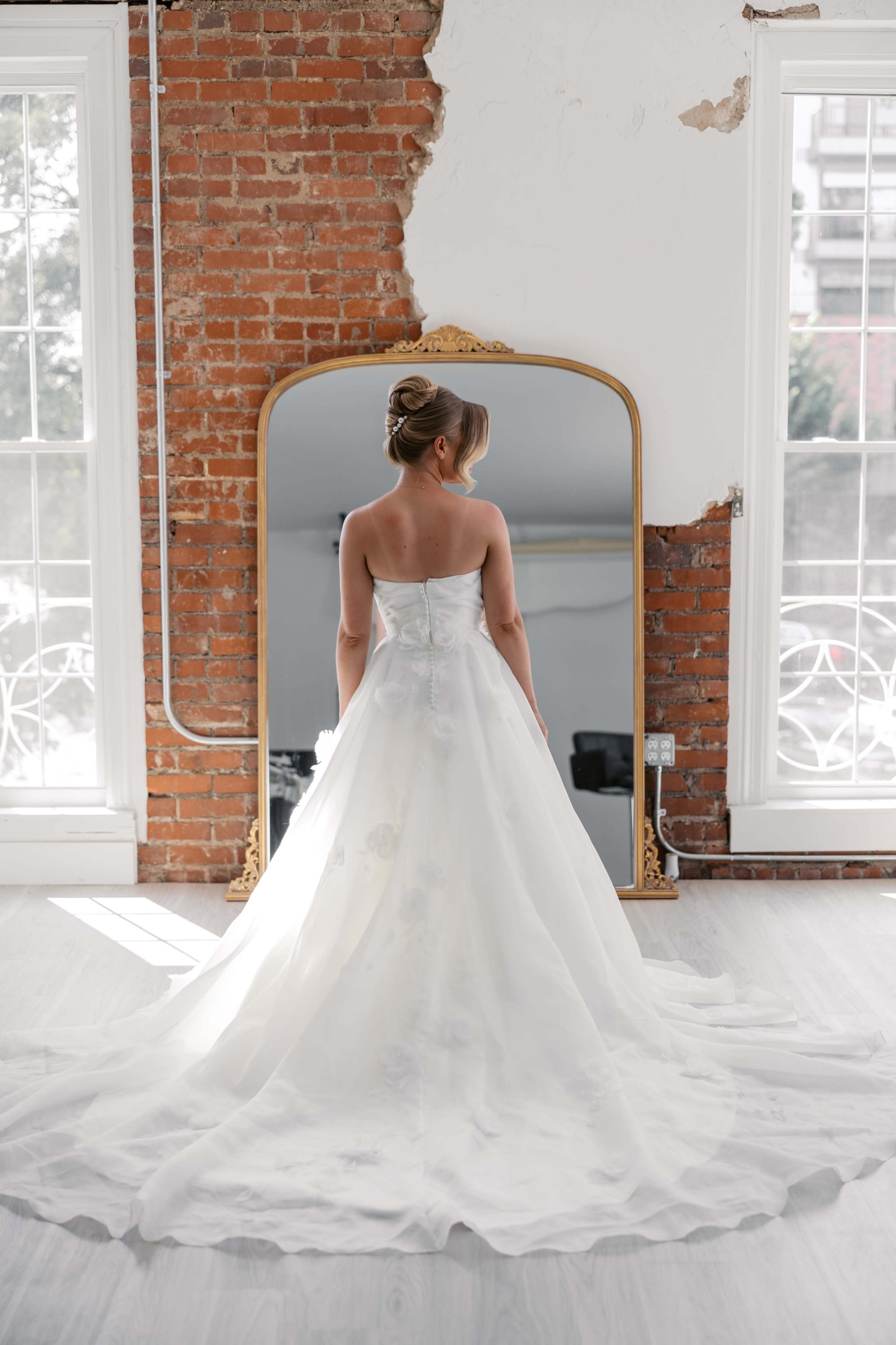 A bride in a white gown stands in front of a large mirror in a room with exposed brick walls and large windows.