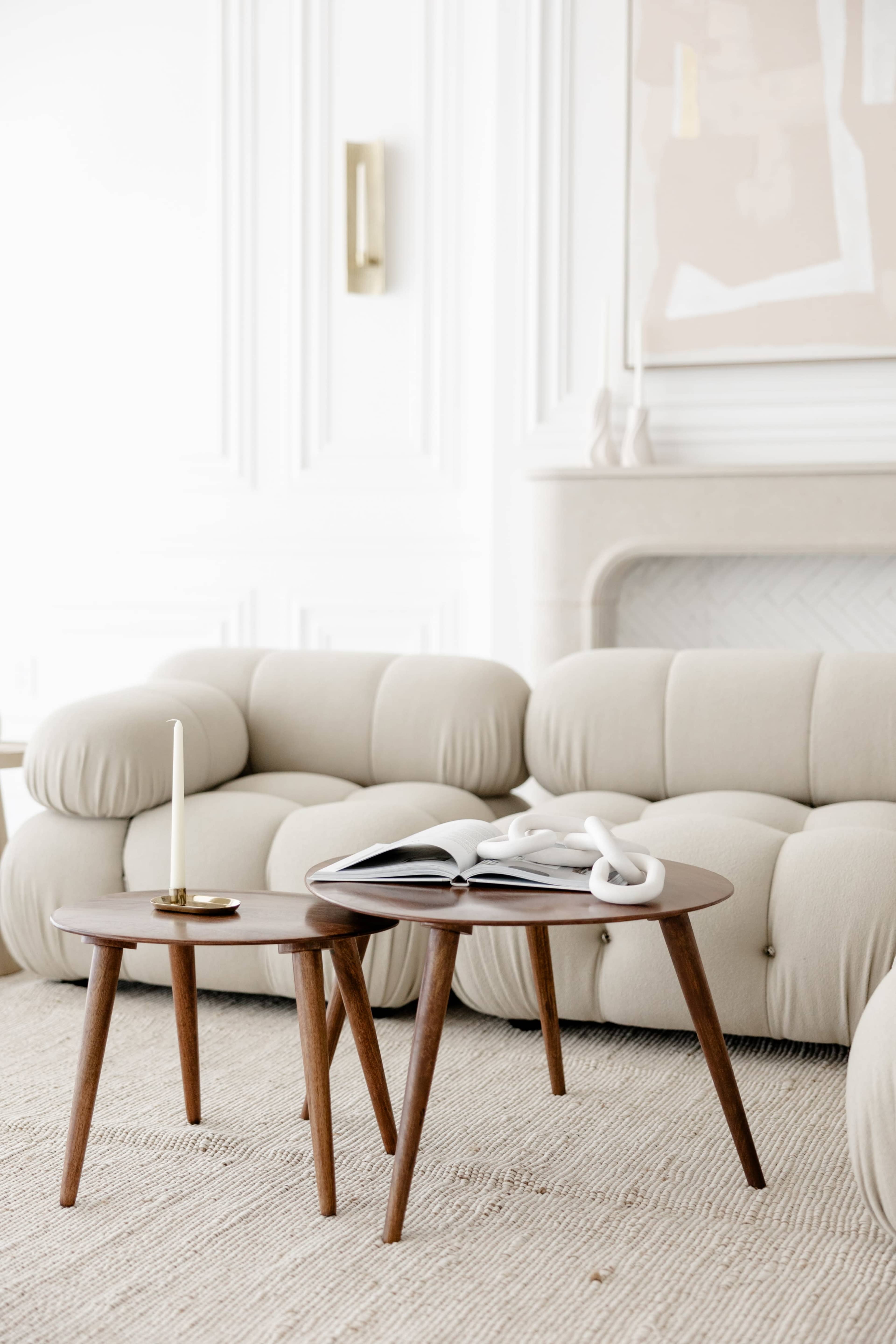A cream-colored, plush sofa rests beside two wooden side tables, one stacked with an open magazine, in a modern, minimalist living room.